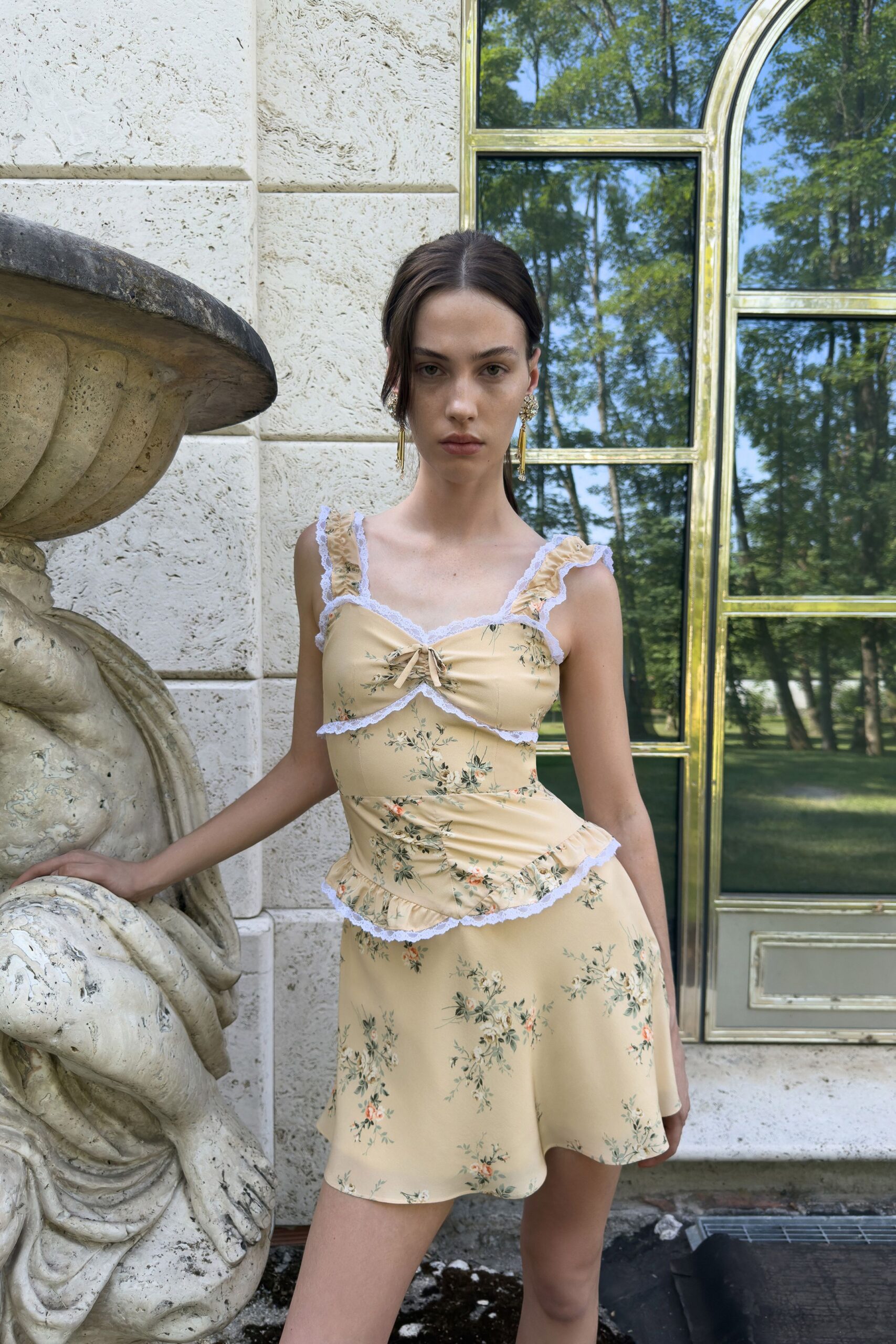 Buttercream satin slip dress scattered with tiny rose sprigs and white lace. Gold cluster earrings with long tassel drops brushing the neck. Garden wedding morning, leaning against cold stone before everyone arrives.