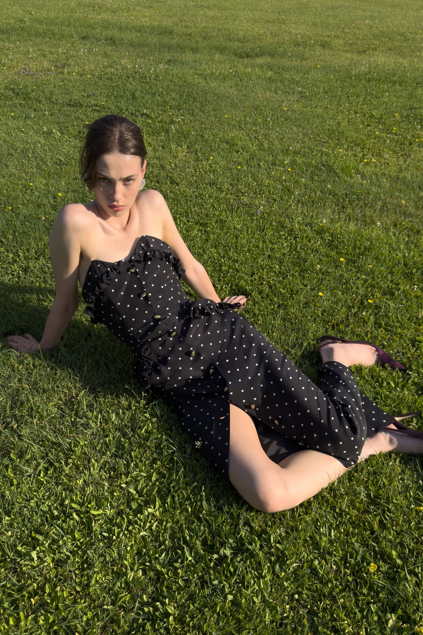 Black strapless polka-dot dress, ruffled neckline and gilt buttons down the split front. Plum pointed pumps with narrow stiletto heels. Sprawled in the grass after a garden party went on too long.