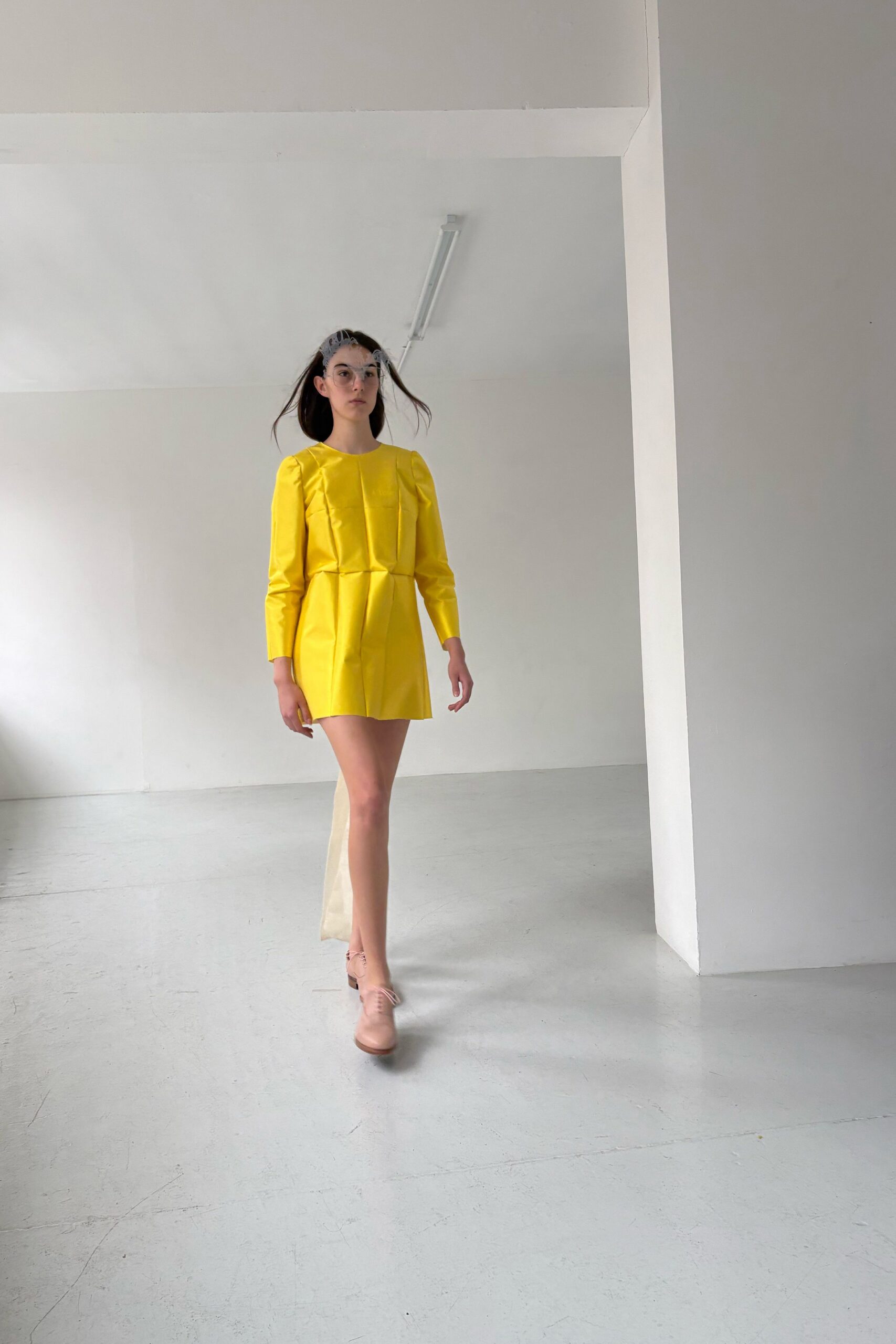 Pale grey lace mask hooked around the temples. Lemon satin mini dress with sharp released pleats and puffed shoulders. Ivory sheer panel falling from the back hem. Blush leather oxfords with a stacked wooden heel. Crossing an empty studio before the paint has fully dried.
