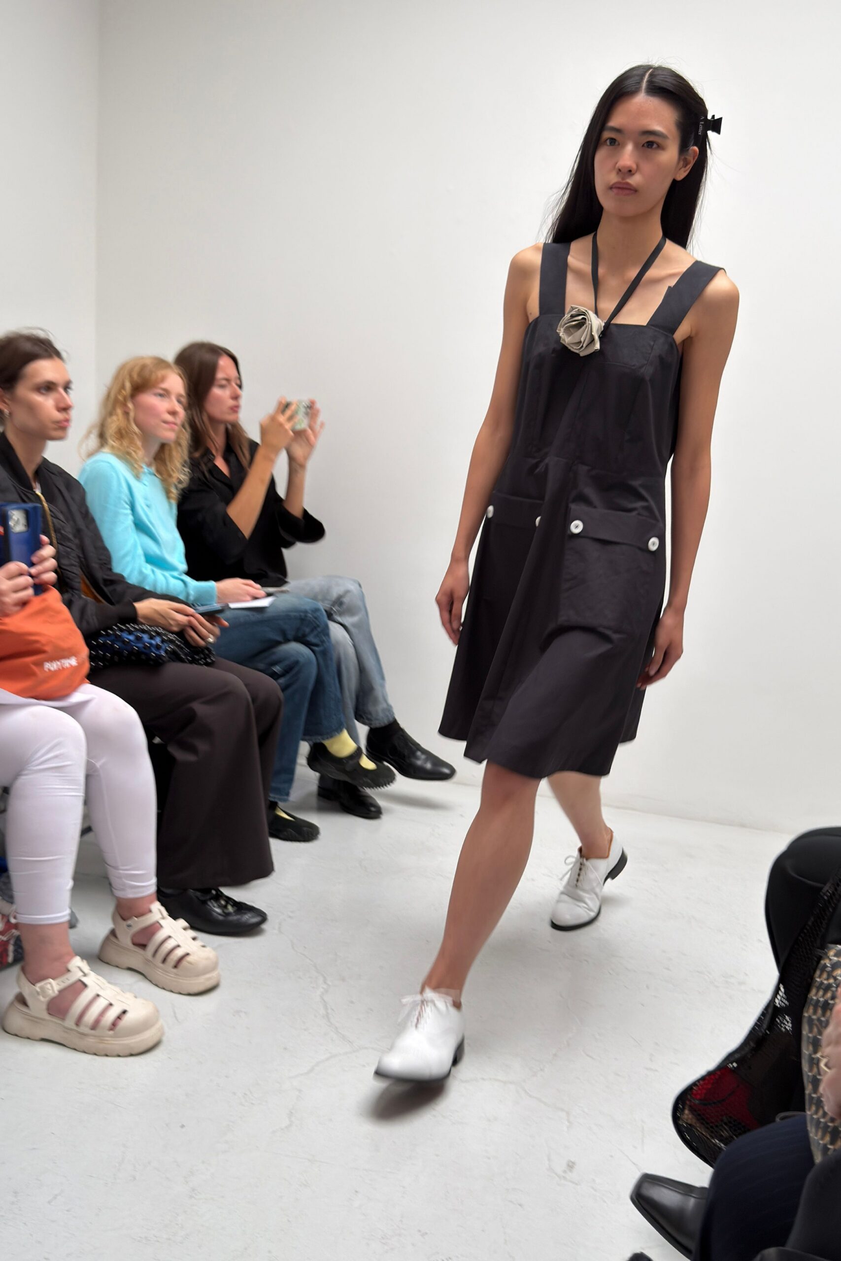 Black cotton pinafore dress with broad straps and white buttoned patch pockets. Taupe fabric rosette hanging from narrow black ties at the chest. White lace-up derbies with fringed tongues brushing the vamp. For walking into a small-town gallery where someone still serves box wine.
