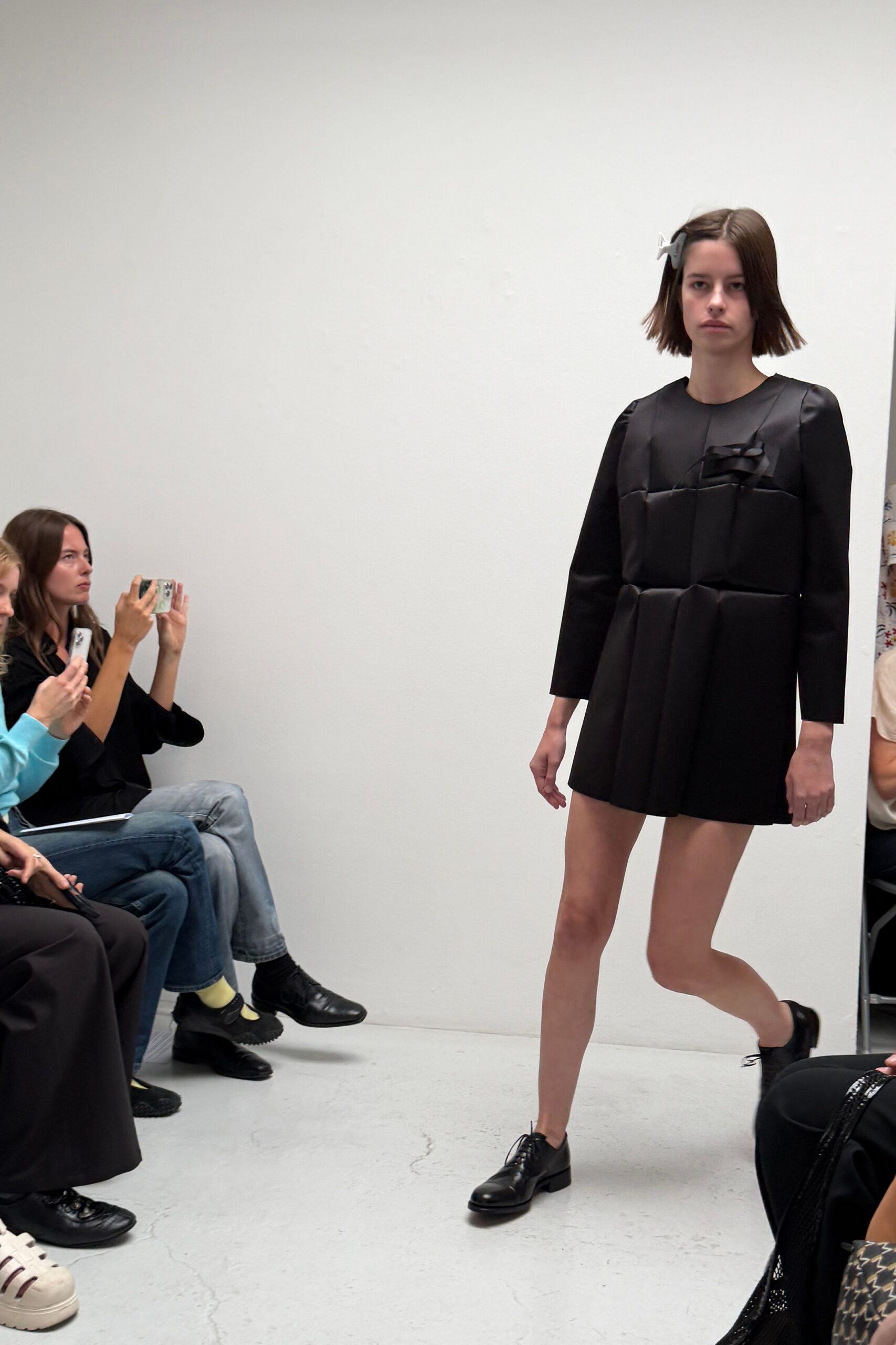 White bow clip pinned flat against a blunt bob. Black satin mini dress built from boxy padded panels, pleats dropped from the waist seam. Black leather derbies with thin round laces. Walking into a school recital still wearing the costume's hardest version.