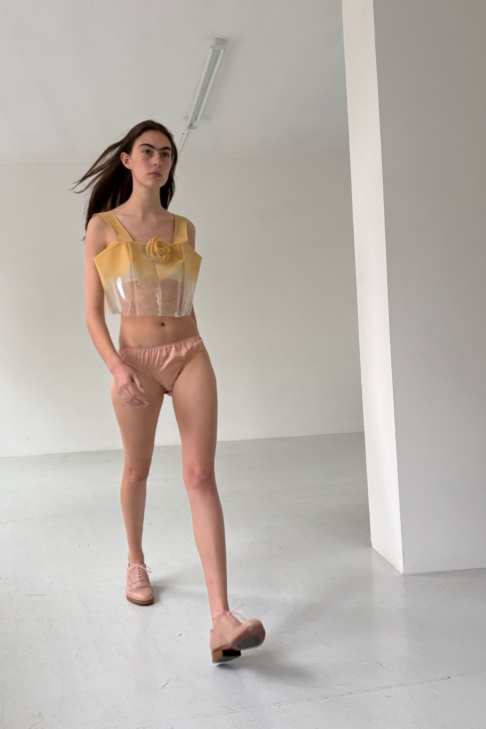 Butter-yellow bustier with a clear vinyl body and rosette at center chest. Blush satin briefs with a softly ruched elastic waist. Pale pink leather oxfords, laces bowing loose over stacked heels. Walking across a photo studio floor still cold from the morning mop.