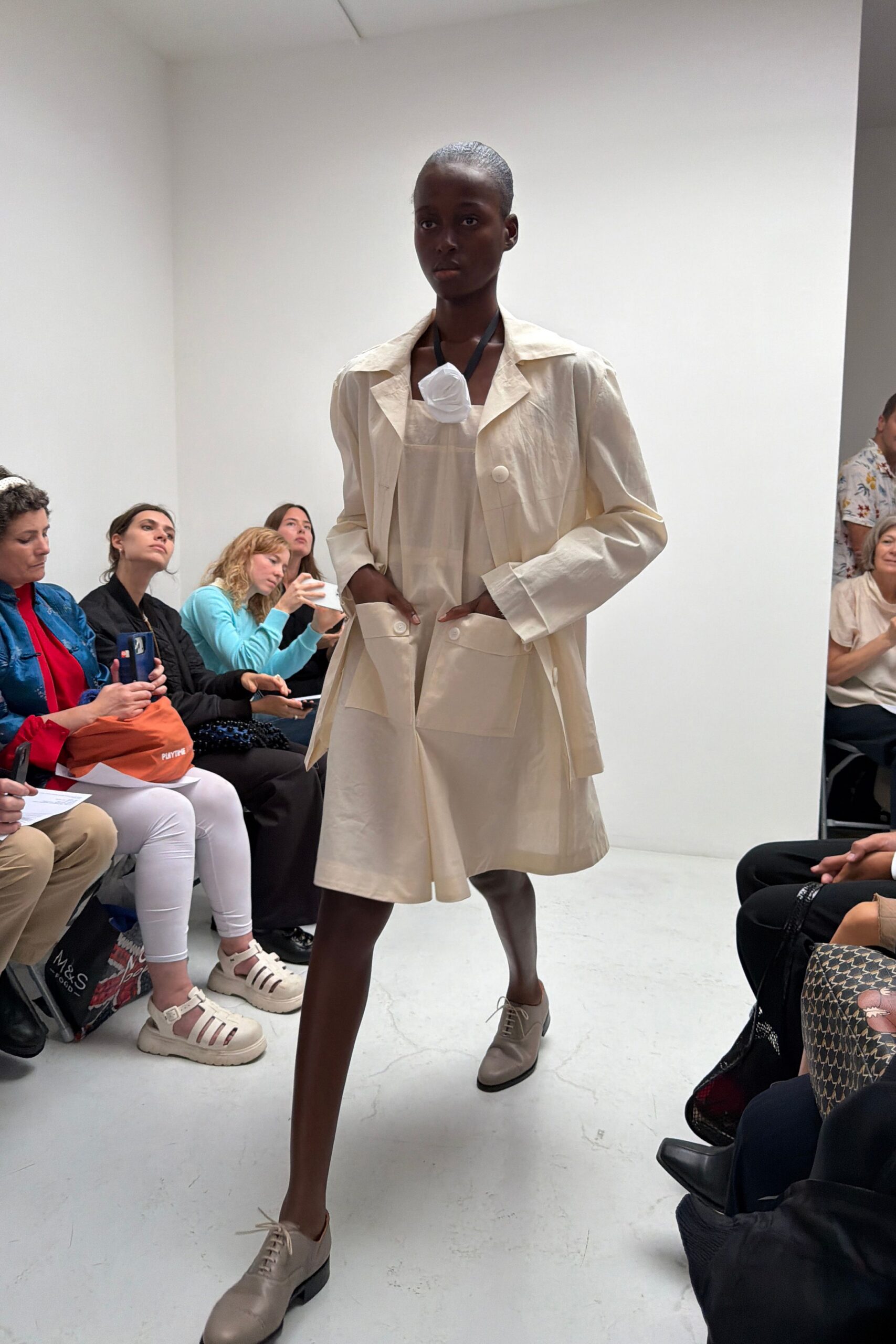 White fabric rosette pendant hanging from a black ribbon. Cream cotton coat with broad lapels and one large covered button. Square-neck cream dress with oversized patch pockets buttoned at the corners. Taupe leather derbies with a low stacked heel. For crossing a museum atrium before the school groups arrive.