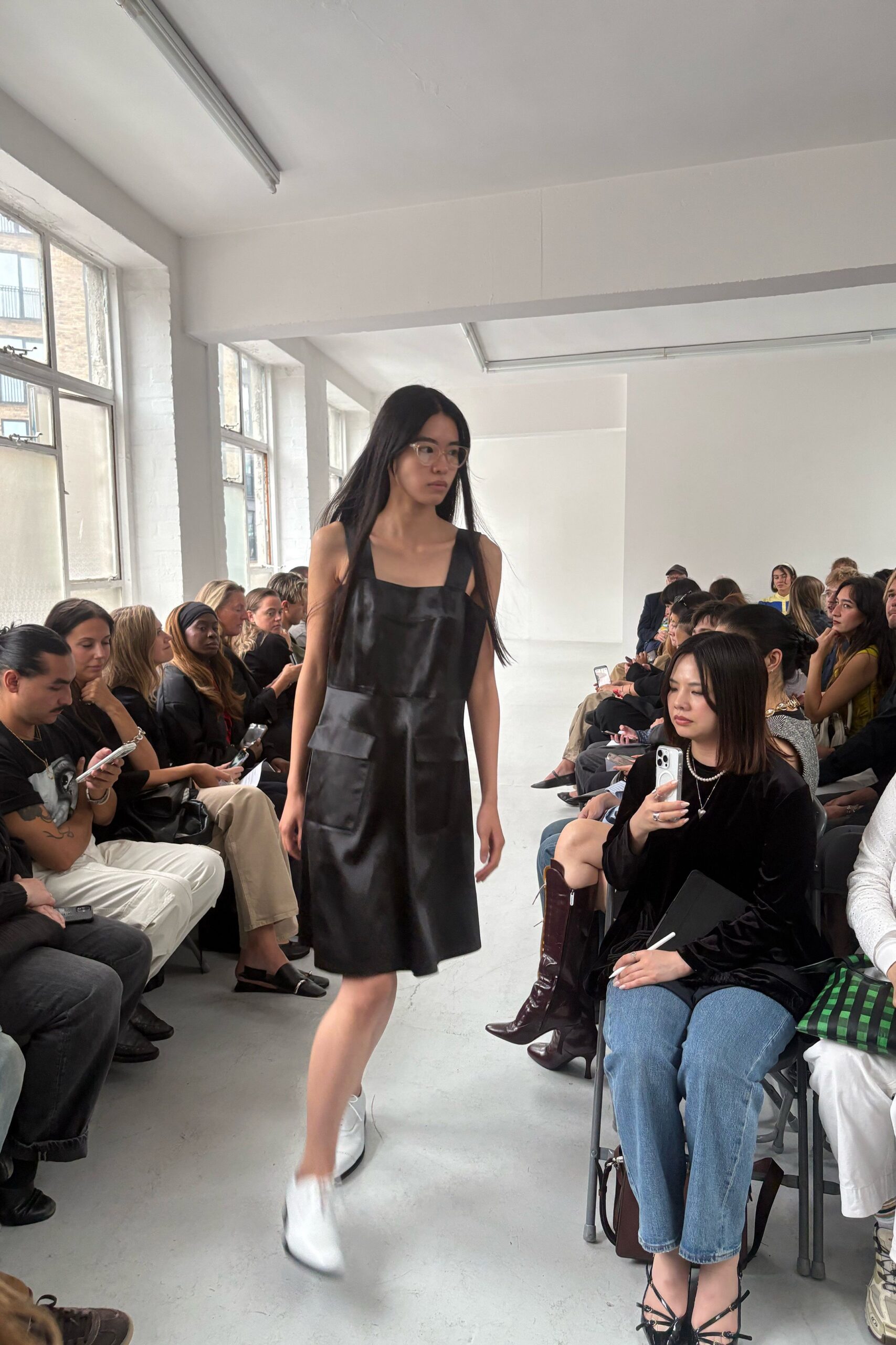 Clear acetate frames with a faintly squared lens. Black coated shift dress, square bib front and one low patch pocket. White leather lace-ups, rounded and plain. Morning in a borrowed studio, windows open to brick dust and traffic.