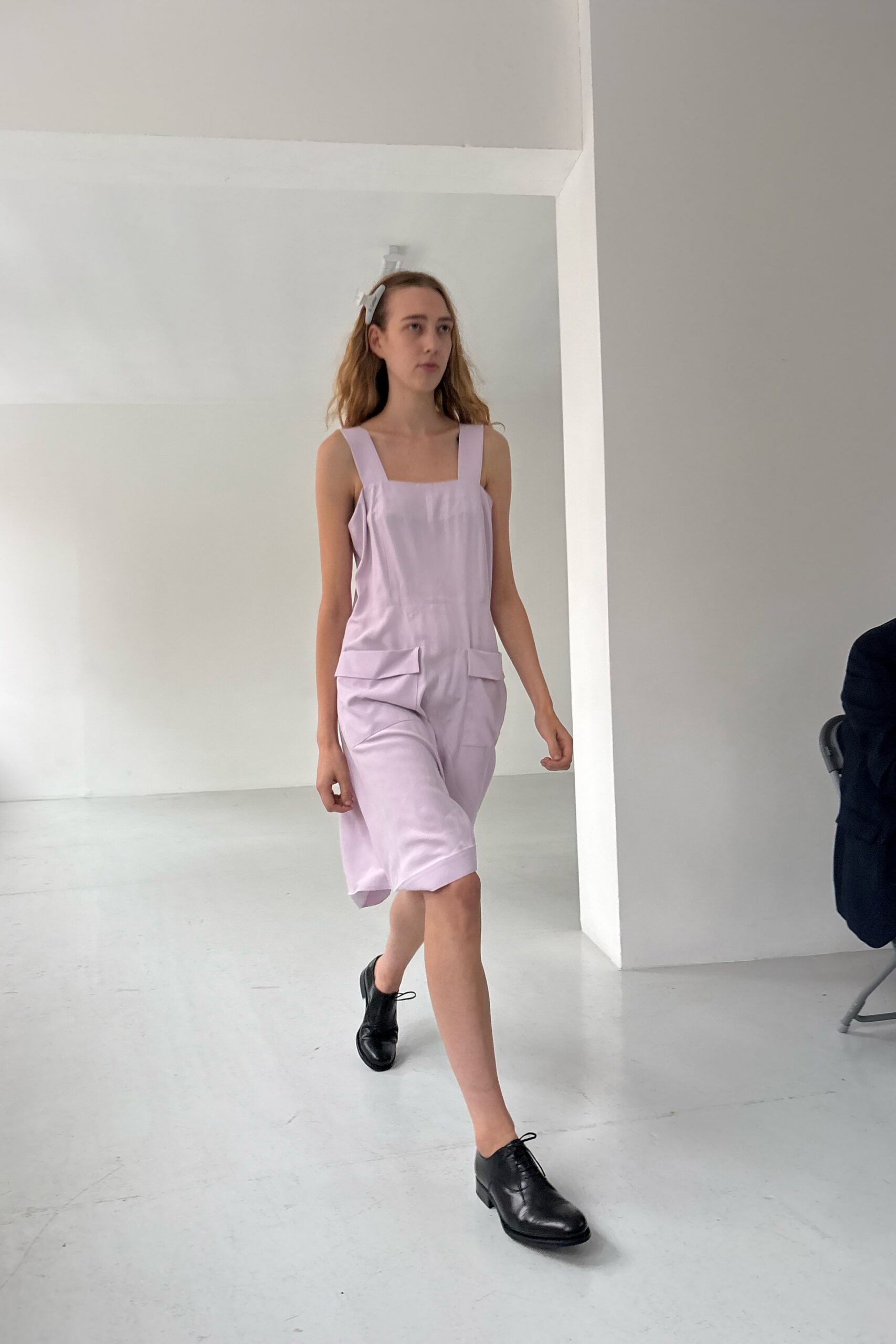 White bow clip pinned above one temple. Pale lilac romper with square bib, dropped flap pockets and cuffed knee hems. Black leather derbies with a low stacked heel. Sunday open house, the rooms still smelling of fresh paint.