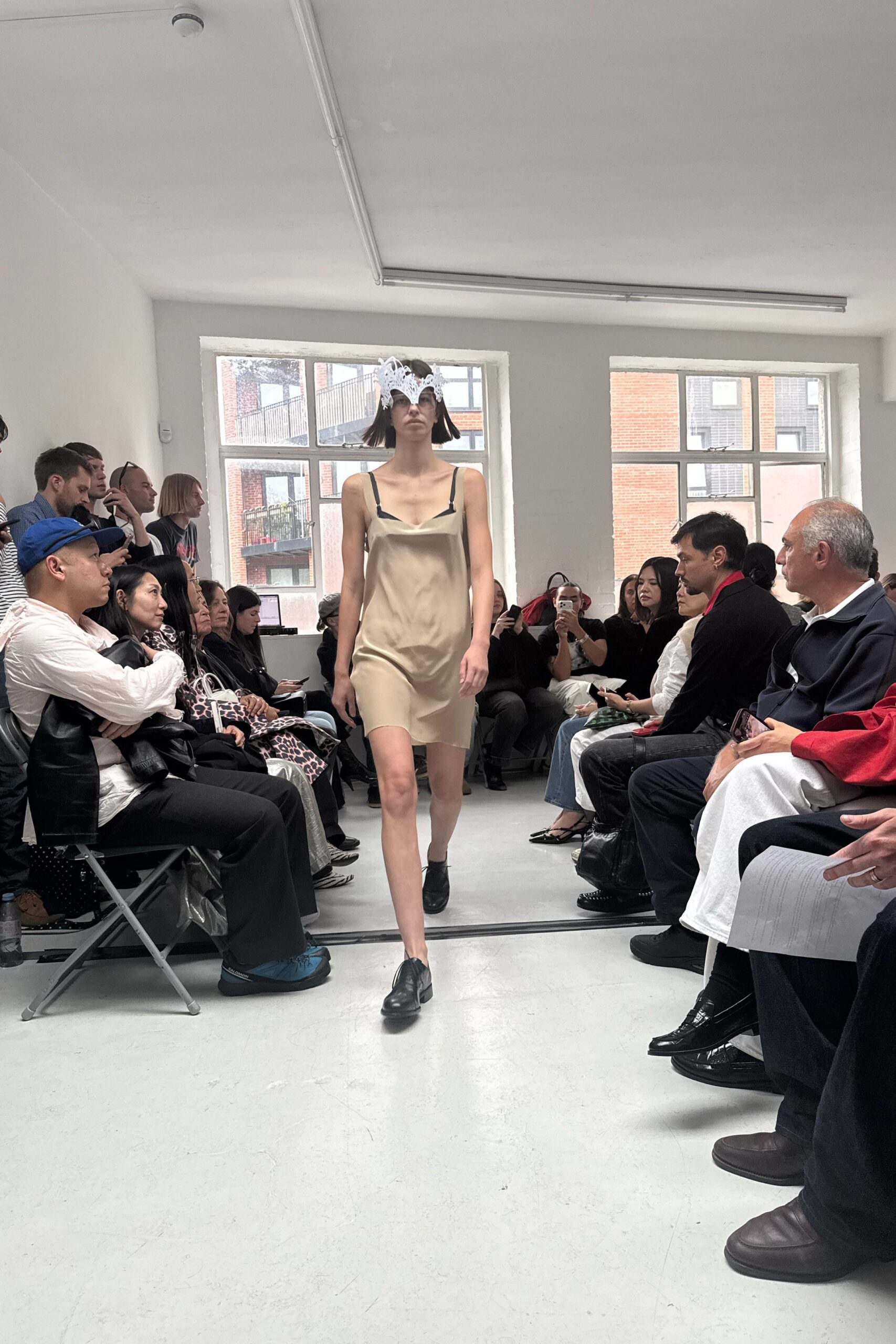 White cutwork mask rising in points over tinted lenses. Champagne satin slip dress, cowl neck sagging between black strap tabs. Black square-toe lace-ups, slightly clunky through the vamp. Morning in a bare studio, the windows still holding last night's rain.