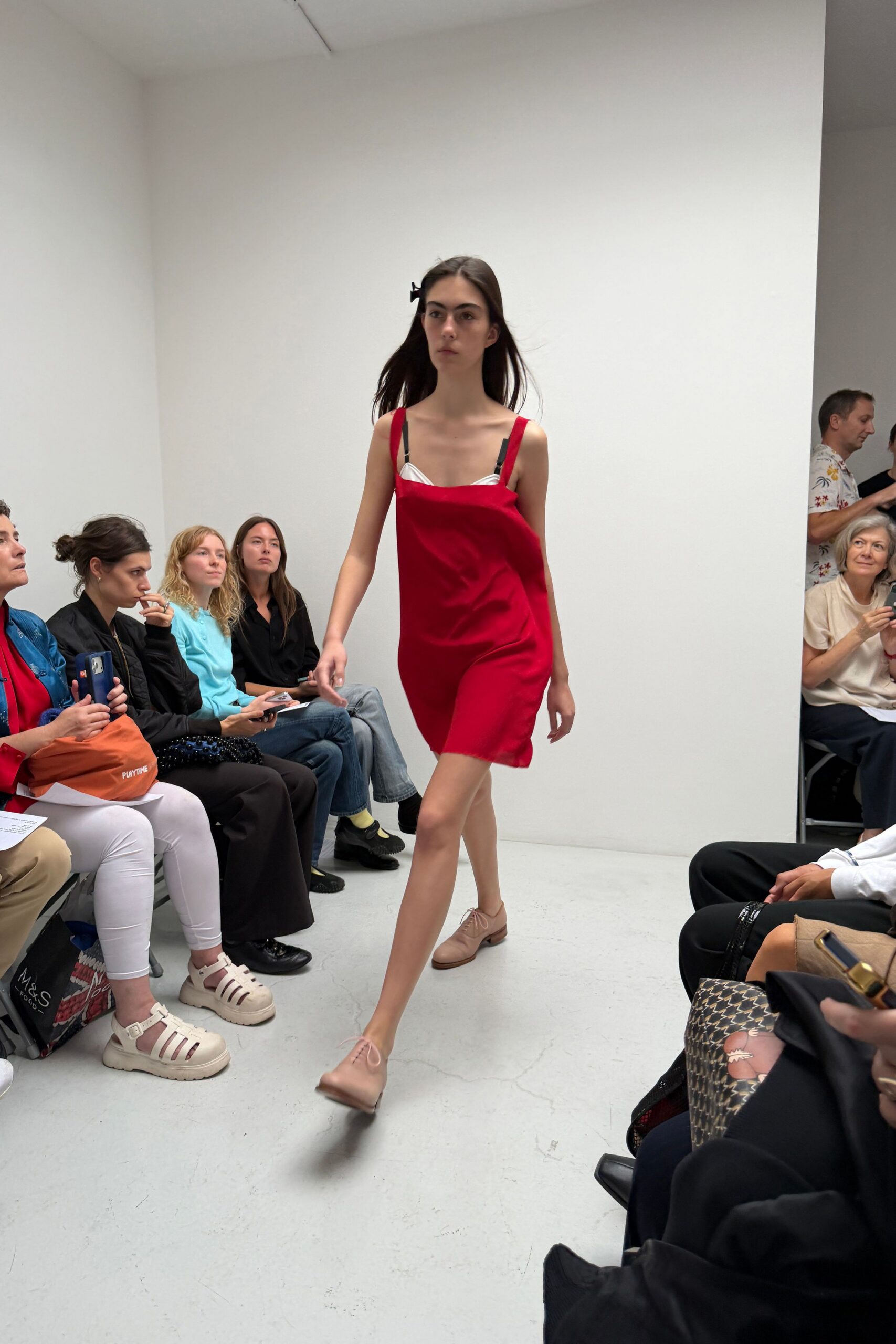 Cherry red satin slip dress, one side sagging lower across the bust. White bra cups peeking above black shoulder straps. Blush leather derbies with slim round laces. Walking into a borrowed apartment with your overnight bag still downstairs.
