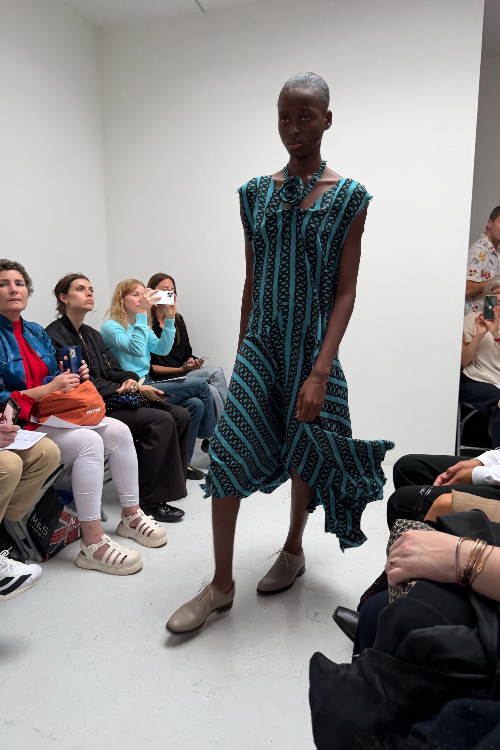 Turquoise sleeveless dress printed with black rope-chain stripes, hem kicked into uneven draped panels. Matching fabric rosette knotted close at the collarbone. Taupe leather derbies with a plain rounded toe. Crossing a tiled courtyard just after the fountain has been turned off.