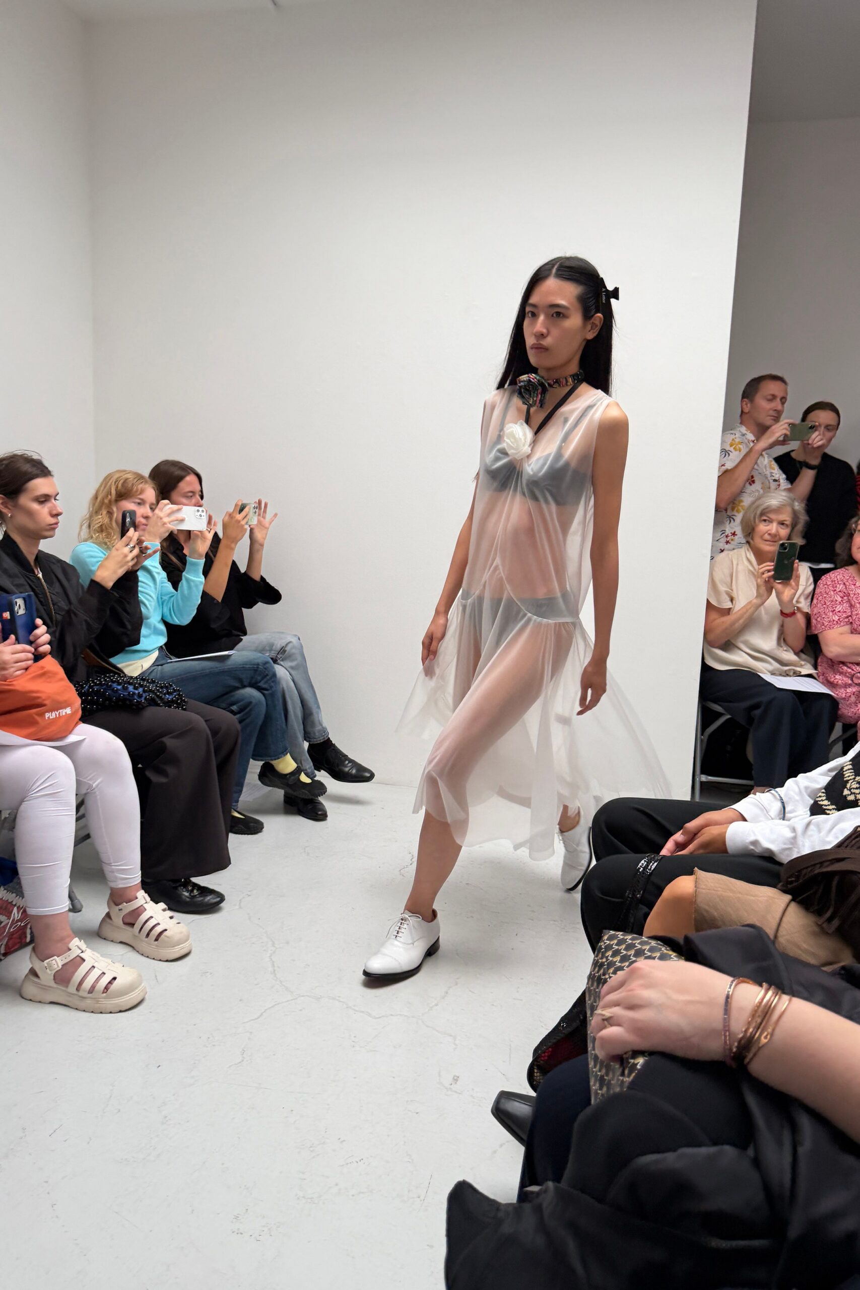 Dark floral scarf tied close, one rosette resting at the throat. Sheer milky chiffon slip dress, dropped seams kicking into uneven godets. Slate triangle bra visible beneath. Matching slate briefs under the gauze. White leather derbies with black-edged soles. Walking across a gallery floor still dusty from the install.