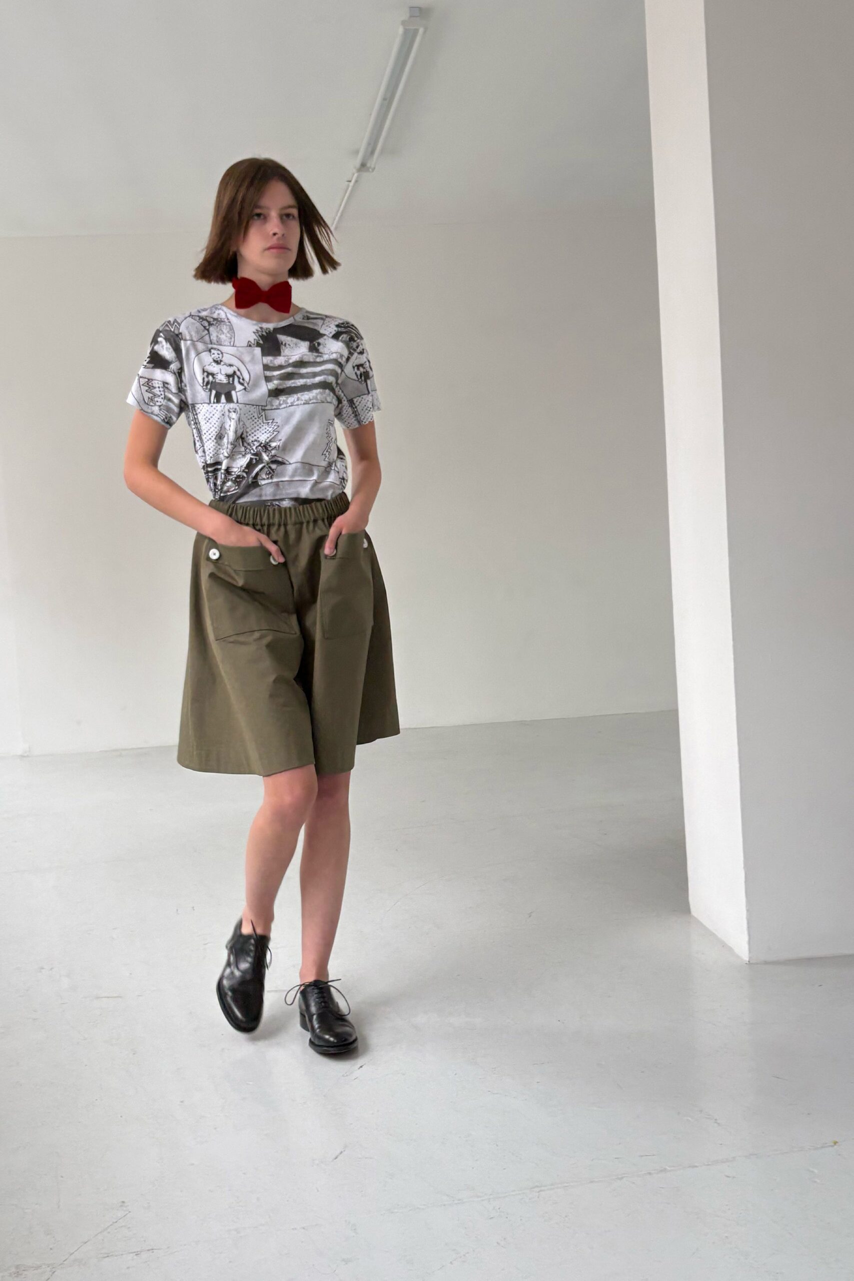 Crimson velvet bow tied close at the throat. Black-and-white comic print tee in slippery satin jersey. Olive cotton shorts cut wide, elastic waist above oversized flap pockets. Black leather oxfords with slightly squared toes. Walking into a municipal art class with charcoal already under your nails.