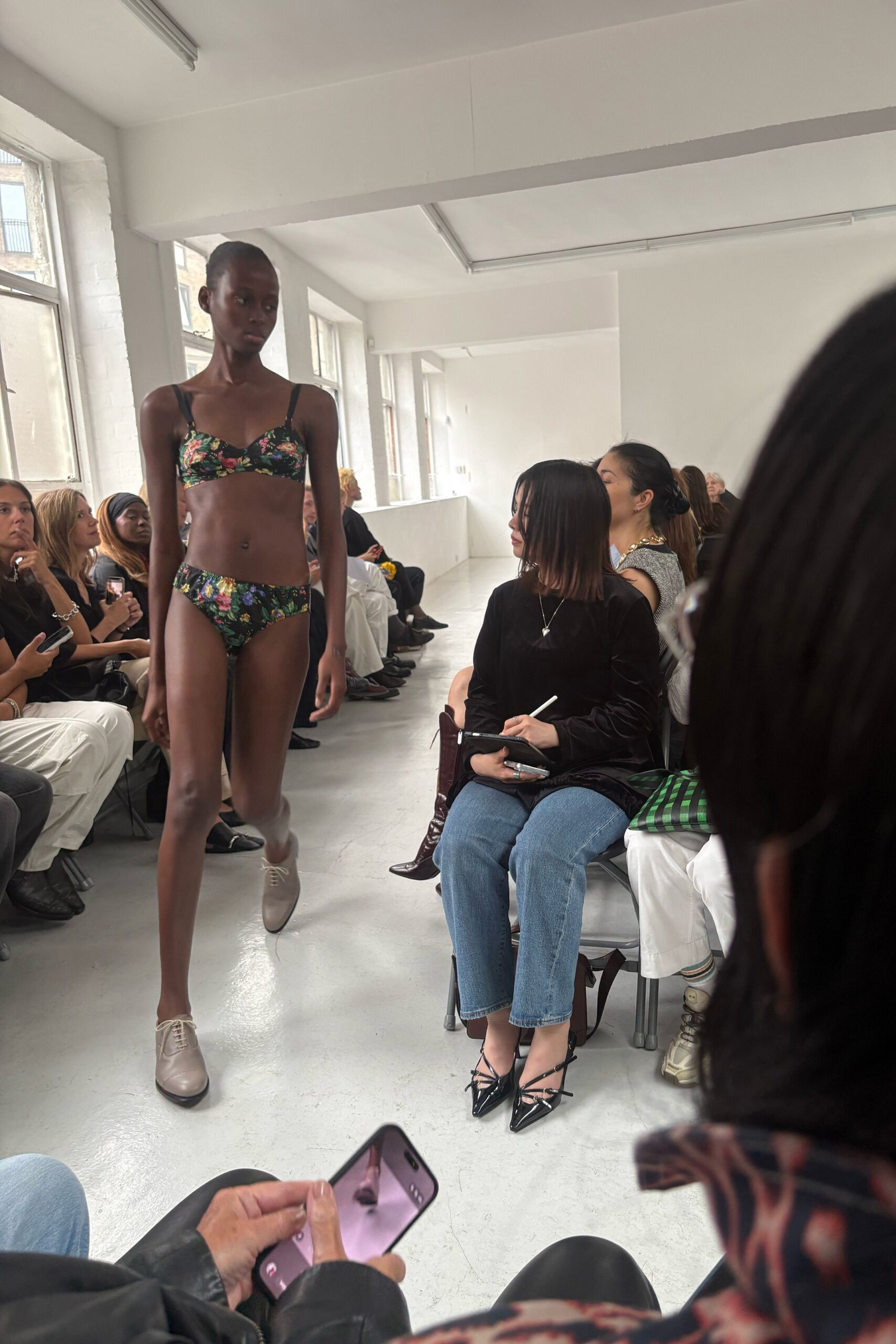 Black floral bra top with gathered cups and narrow shoulder straps. Matching low-rise briefs in glossy dark floral stretch. Pale greige leather derbies with blunt black-edged soles. Walking back from a hotel pool through the corridor in borrowed shoes.