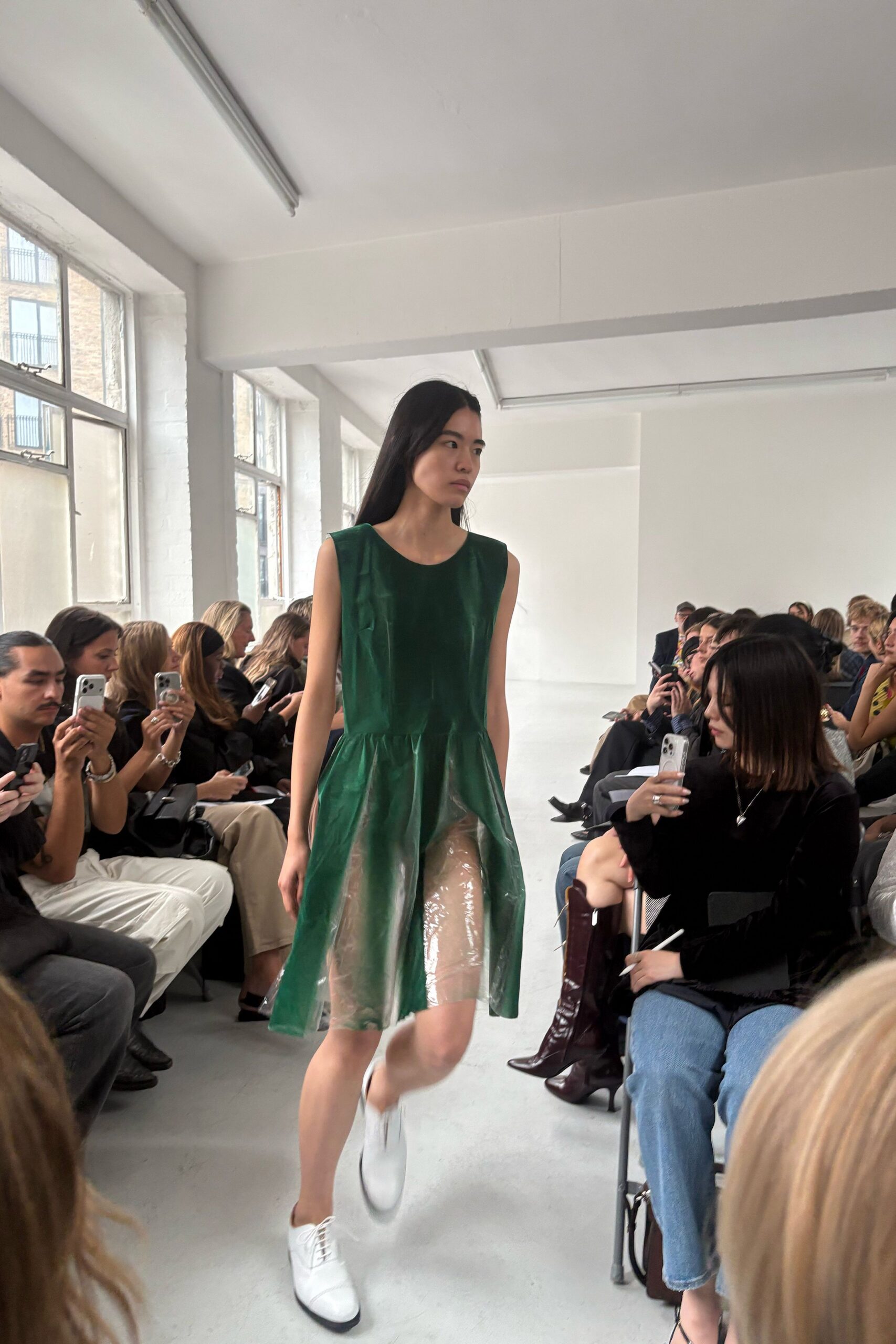 Bottle-green sleeveless dress with a glossy clear vinyl skirt dropped from the waist. White lace-up sneakers with black-edged soles. Walking into an empty conservatory just after the plants were watered.