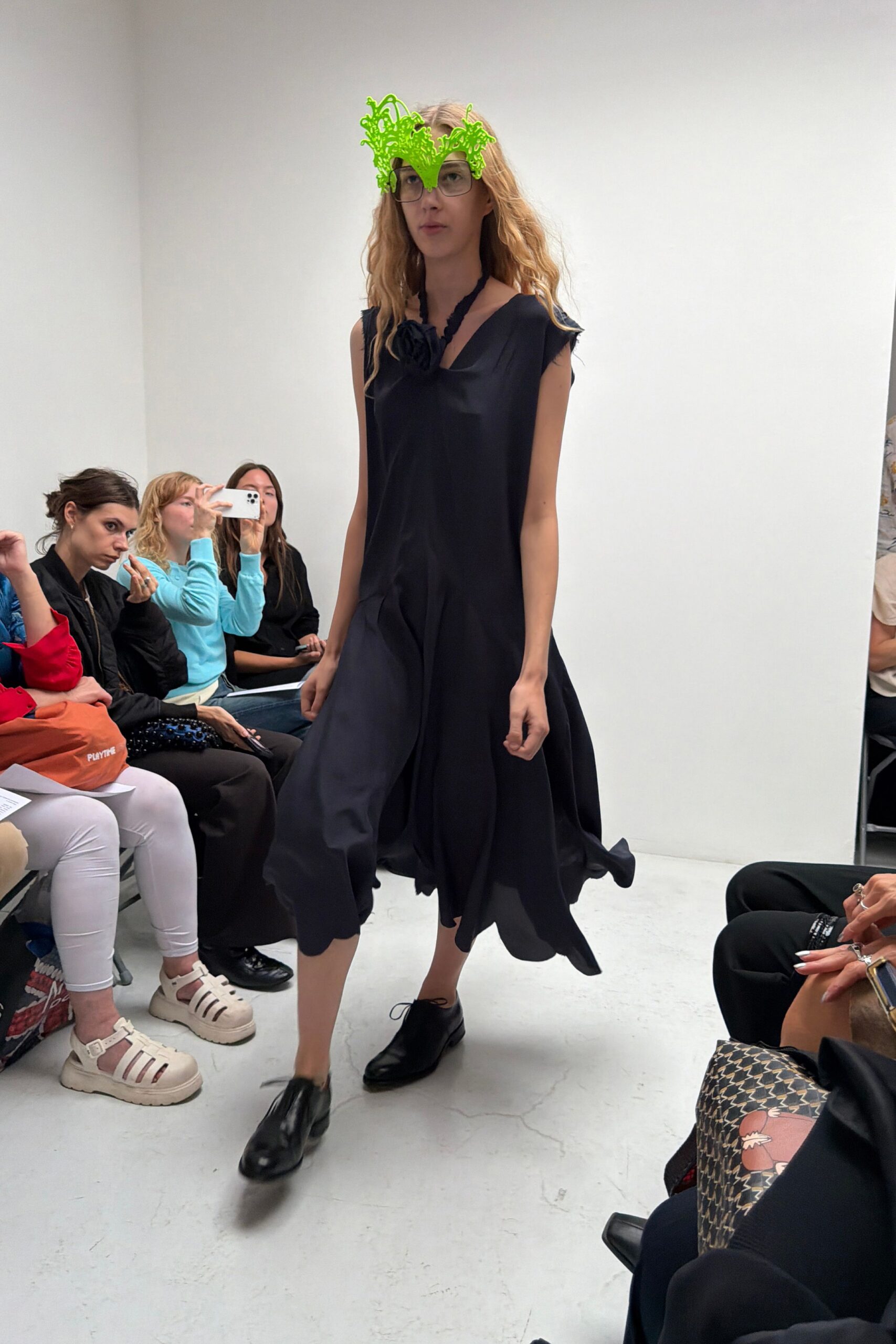 Oversized square glasses topped with a neon green squiggle visor. Black sateen midi dress, cut loose with a drooping rosette neckline. Black leather lace-ups, one toe catching the light. Walking into a student crit with wet hair and one wild idea left.