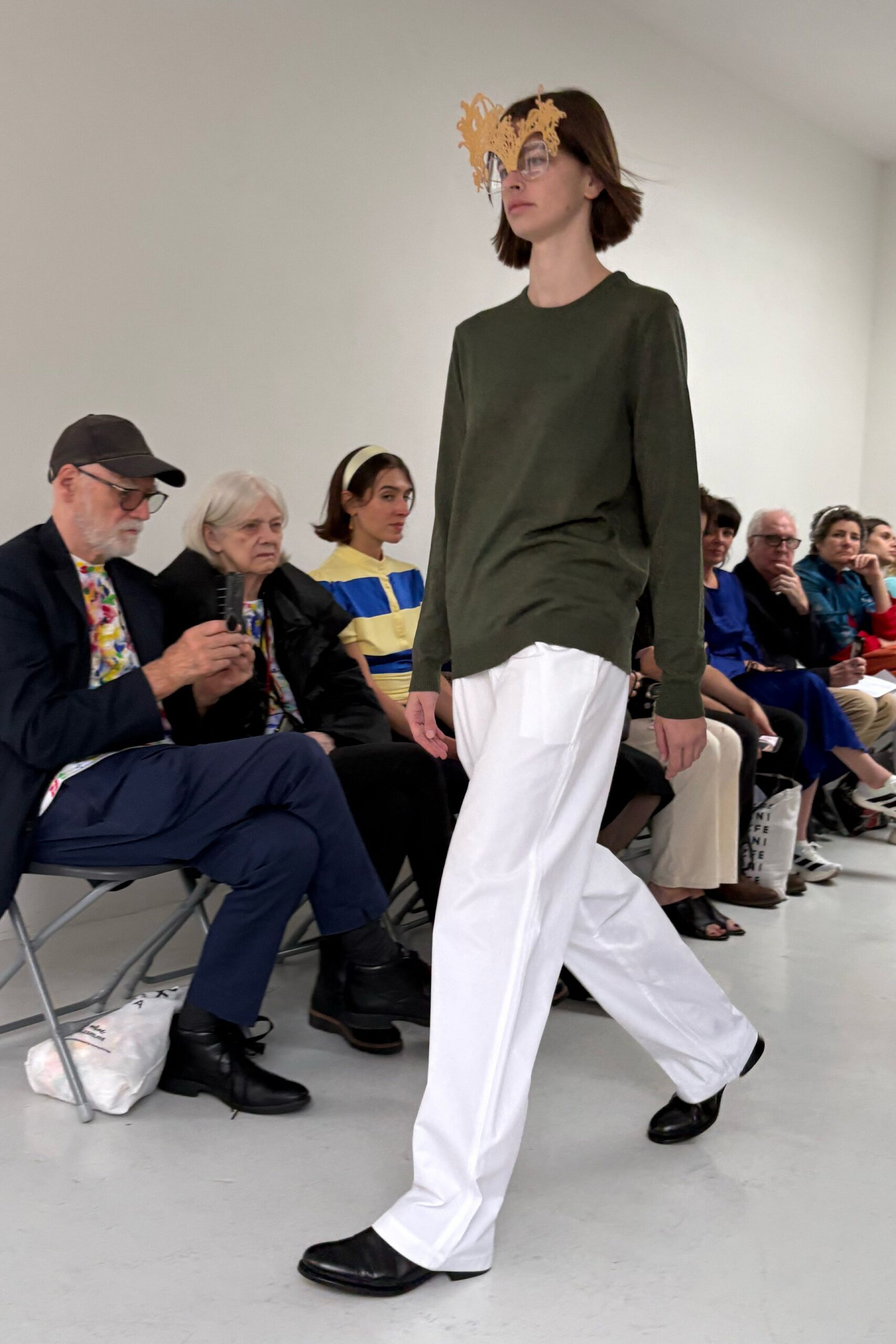 Clear shield glasses topped with a jagged mustard filigree brow. Moss fine-knit crewneck hanging long, one side caught into the hip. White wide-leg trousers in crisp cotton, creased hard down front. Black square-toe flats with a low stacked heel. Walking a white corridor while the paper invitation softens in your hand.