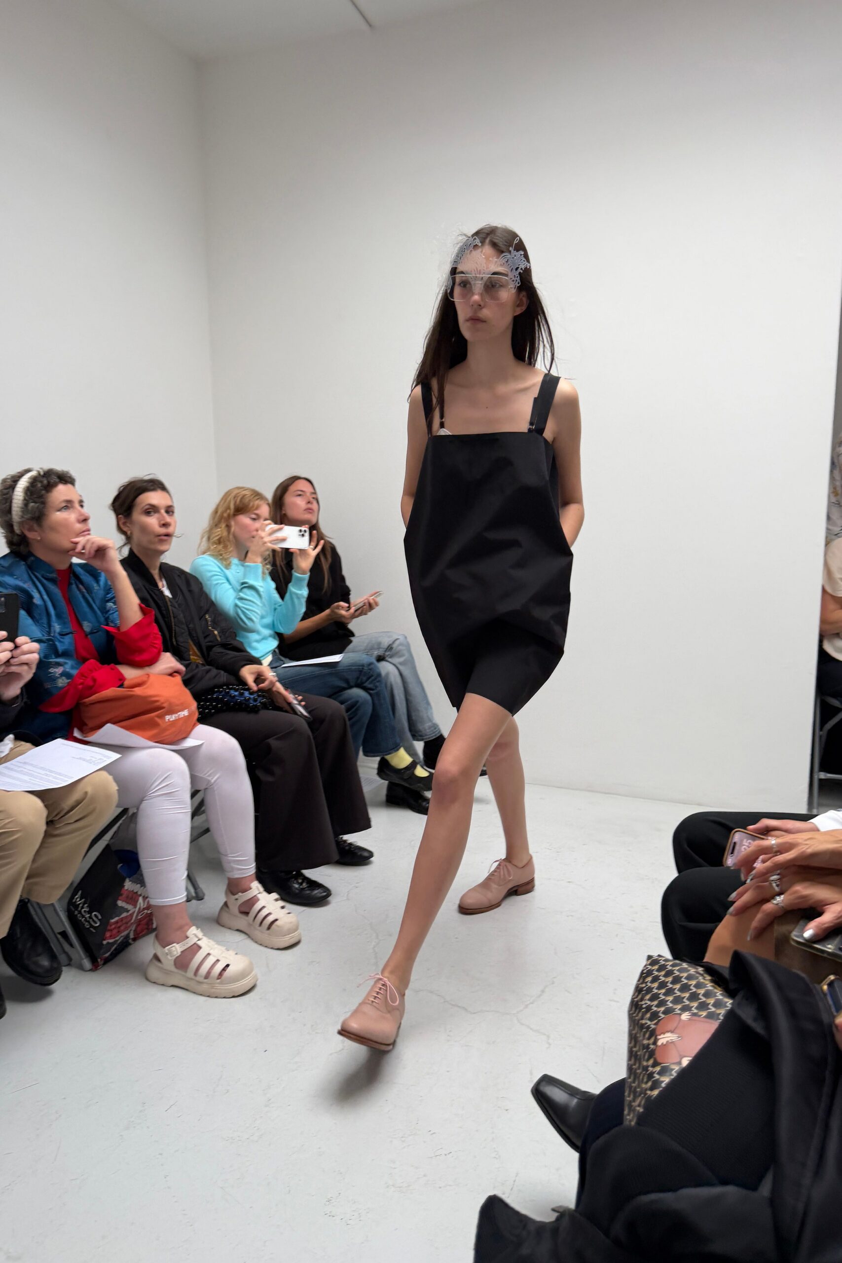 Clear shield glasses edged with white lacework curling over the brow. Black structured mini dress puffed outward, bib front hung from clipped straps. Cycling shorts peeking below the uneven hem. Blush leather derbies with long loose laces. Walking into a bridal fitting with your school shoes still on.