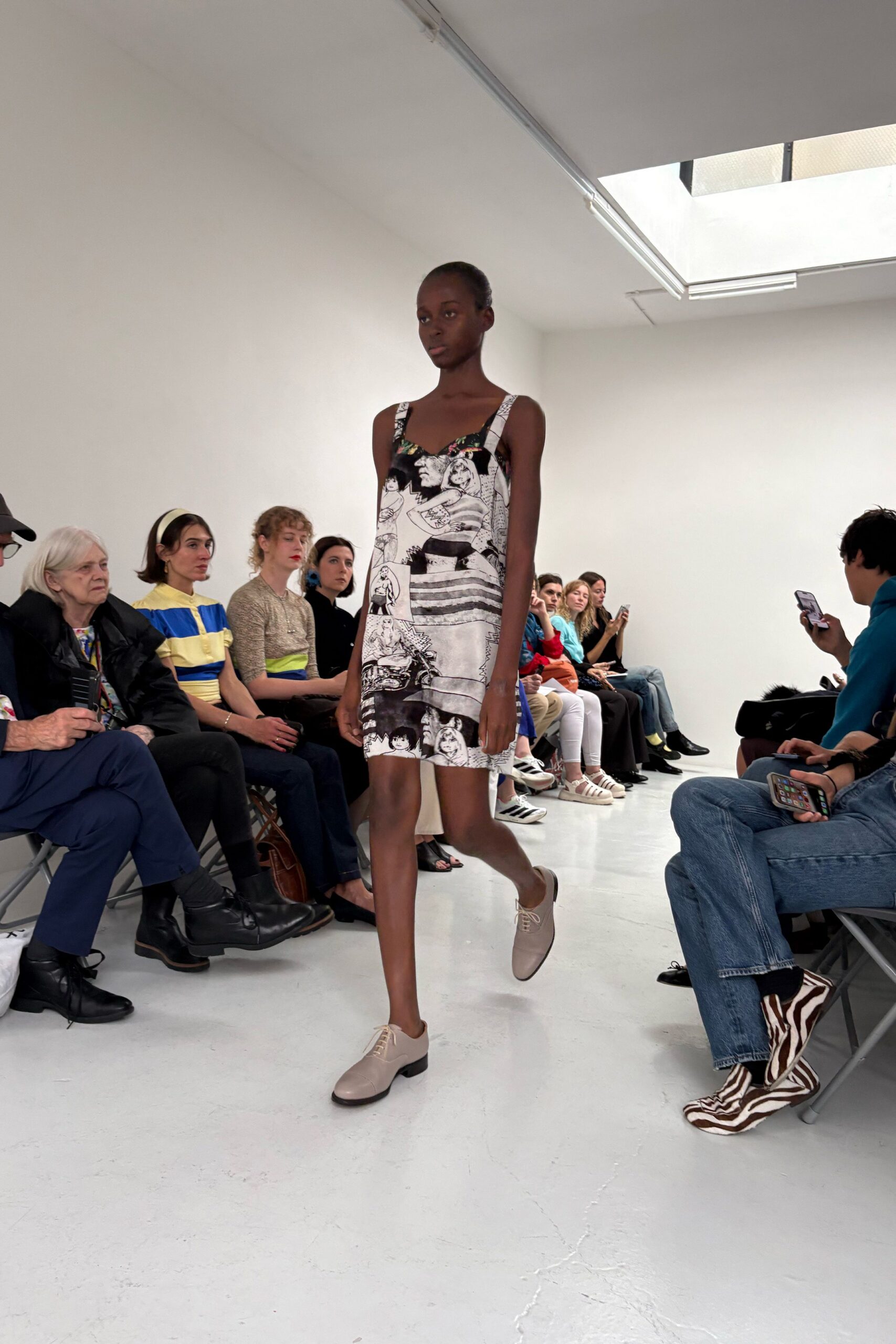 Black-and-white comic-panel slip dress with broad printed straps and a shallow sweetheart neckline. Pale greige leather derbies with slim laces and stacked black heels. Waiting out a summer downpour in a gallery district copy shop.