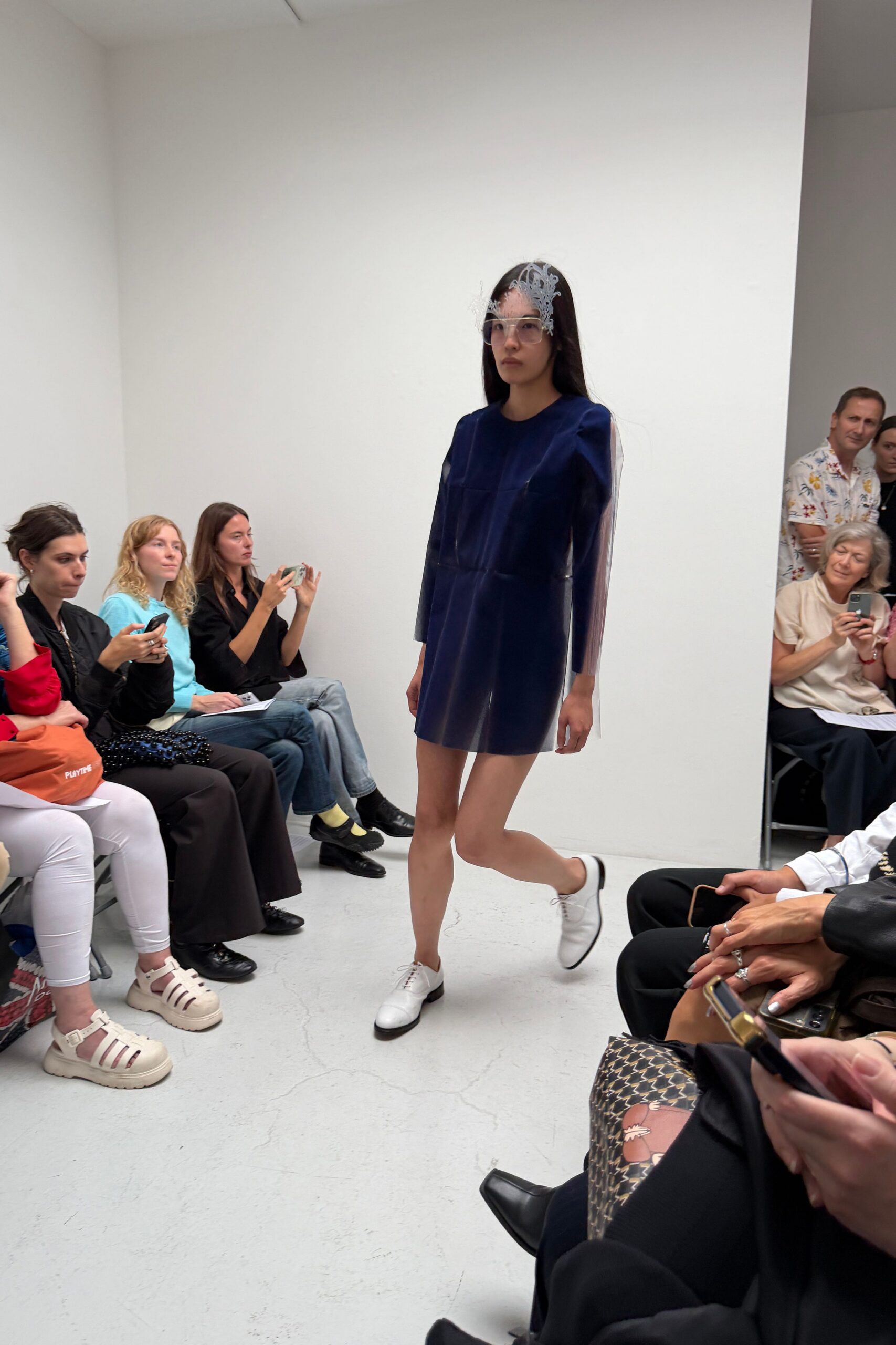 White lacework veil clipped over one eye, feathery at the edge. Oversized clear frames with a blunt rectangular front. Inky shift dress in glossy bonded panels, sheer plastic sleeves hanging straight. White leather derbies with black-tipped soles. Museum atrium at noon, footsteps bouncing off the white walls.