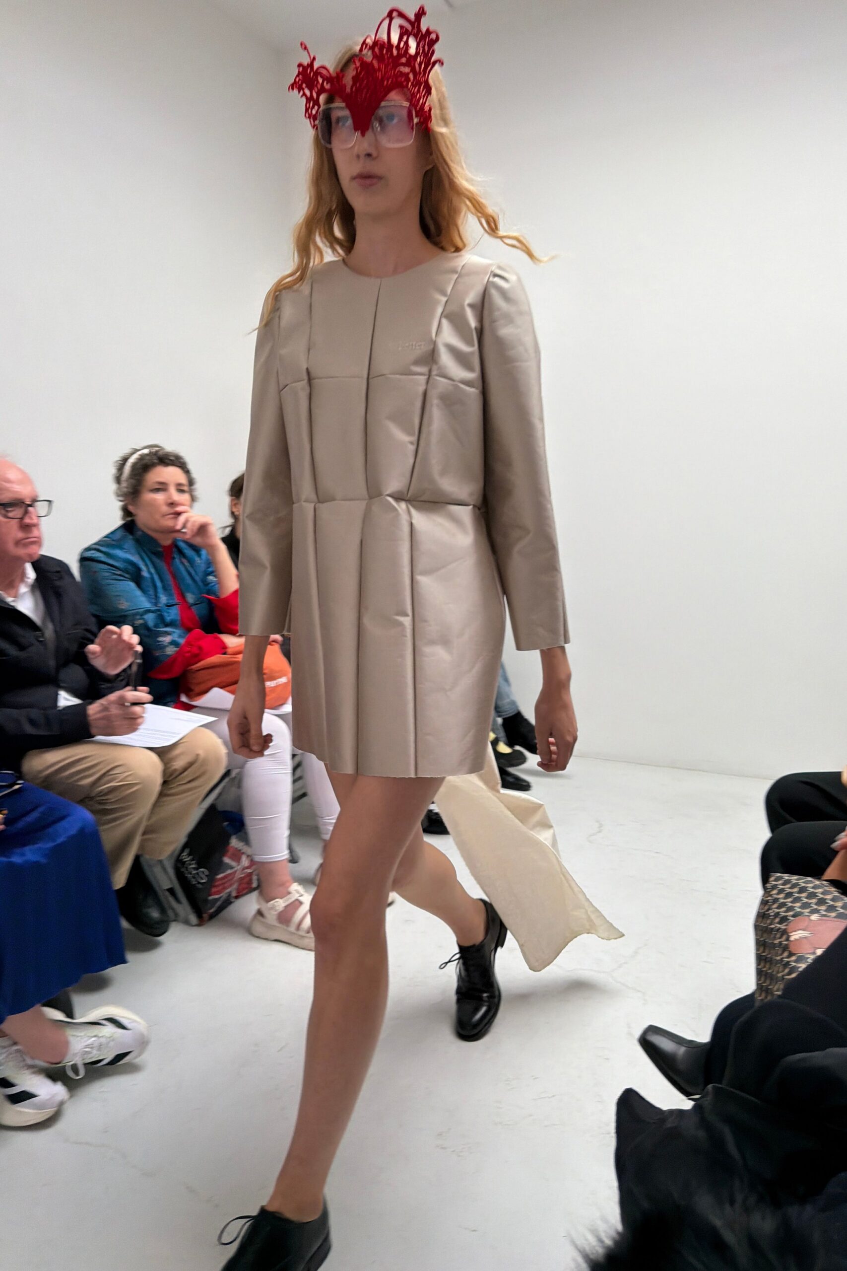 Oversized translucent frames crowned with a red looped wire flourish. Pale putty shift dress in crisp bonded cloth, folded into boxy vertical panels. Long ivory panel dragging from the back hem. Black leather derbies with thin laces. Walking into a municipal wedding hall with the wrapping paper still attached.