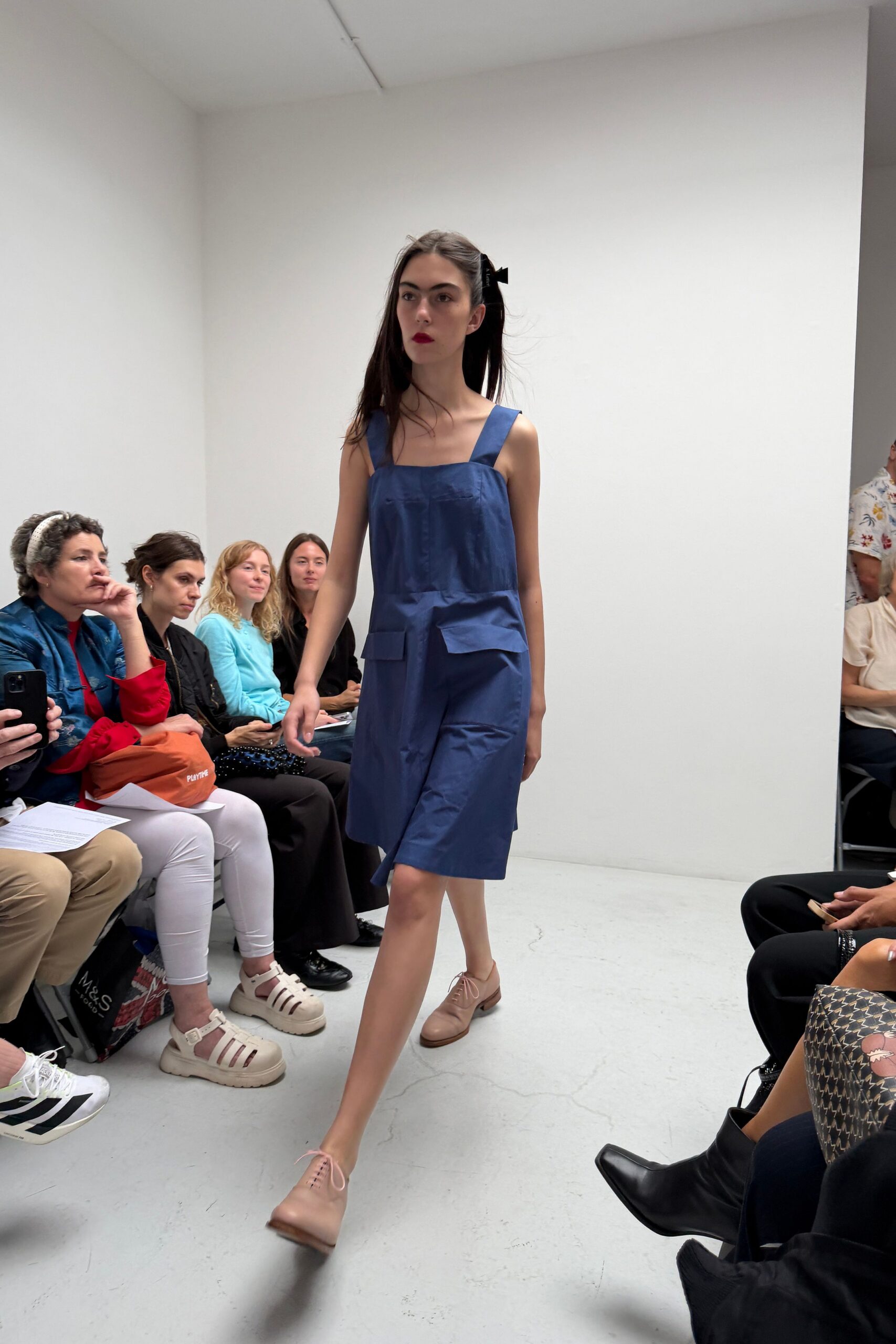 Ink-blue cotton pinafore dress with broad straps and squared patch pockets. Blush leather derbies, throat left bare where the laces start. Black ribbon bow clipped at one side. Walking into a school hall repurposed for a summer art fair.