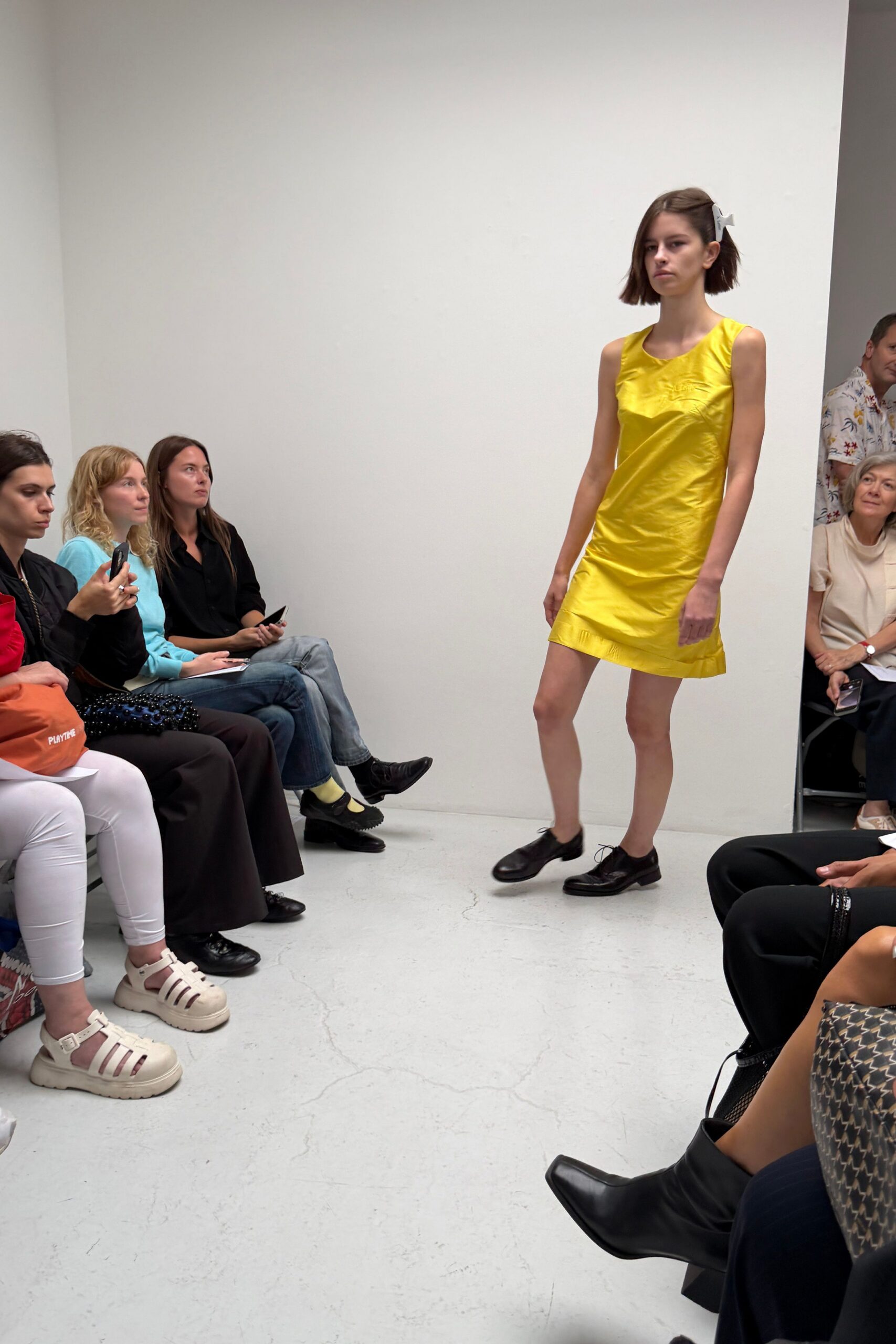 Lemon satin shift dress, one shoulder slightly dropped and hem lightly folded. White bow clip pinned behind the bob. Dark leather derbies with thin laces and rubbed toes. Walking into a church hall wedding before the cake is cut.