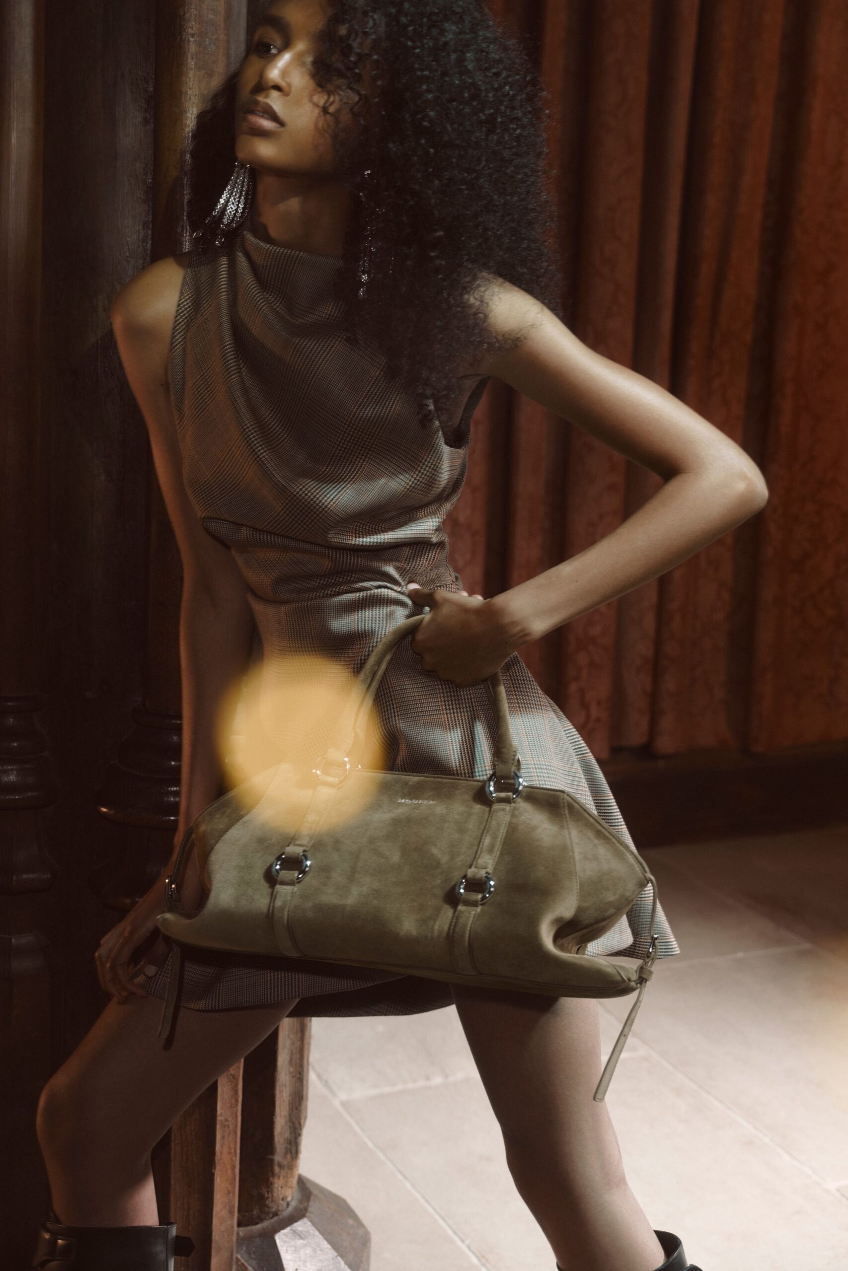 Long crystal tassel earrings brushing the jaw. Sleeveless taupe micro-check dress with a liquid cowl neck and flared hem. Olive suede holdall with silver O-rings and one strap end hanging free. Black leather ankle boots. Waiting in a paneled hotel corridor before the wedding guests spill out.