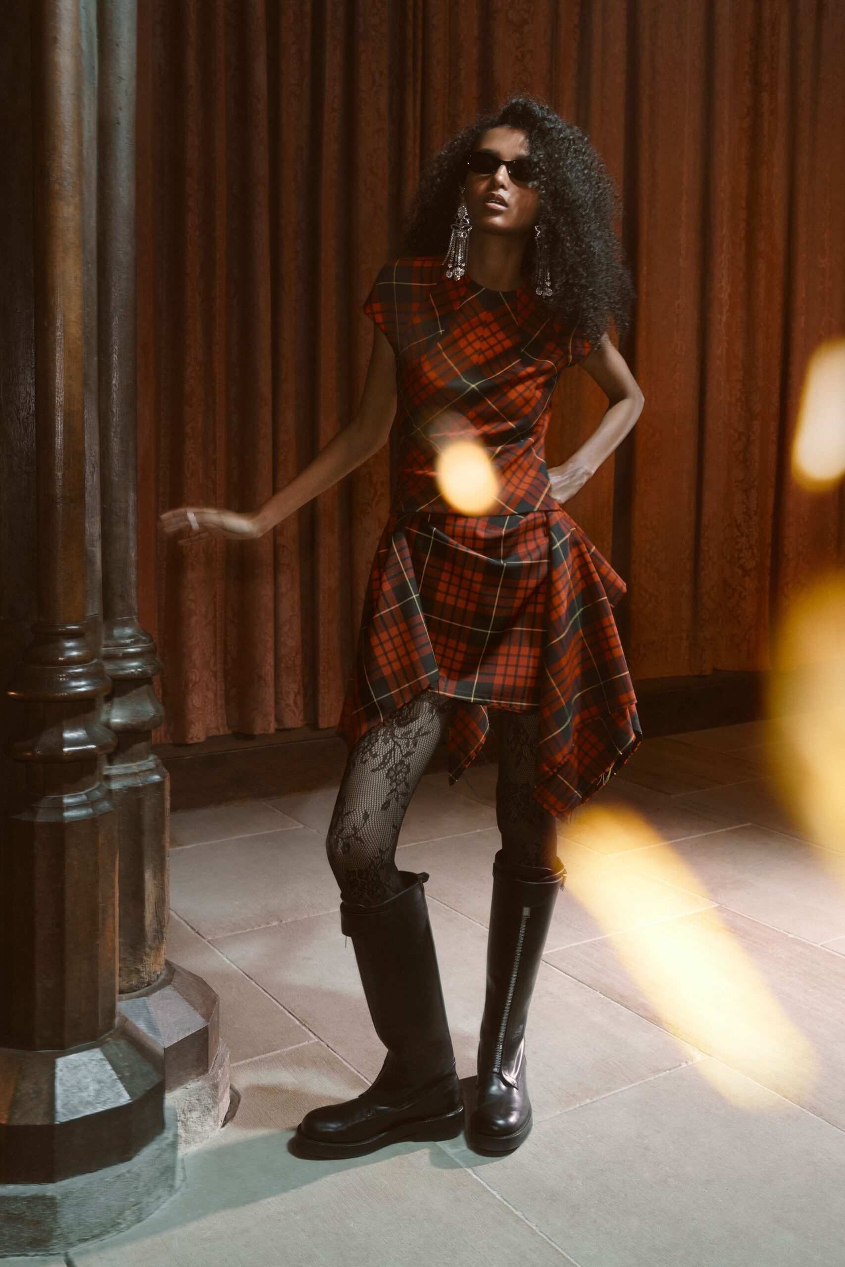 Narrow black sunglasses. Crystal chandelier drops grazing the shoulders. Red tartan dress with cap sleeves and an uneven handkerchief hem. Black lace tights, floral scrollwork climbing one thigh. Knee-high black leather boots with front zip seams. Crossing a hotel ballroom before the wedding band starts.