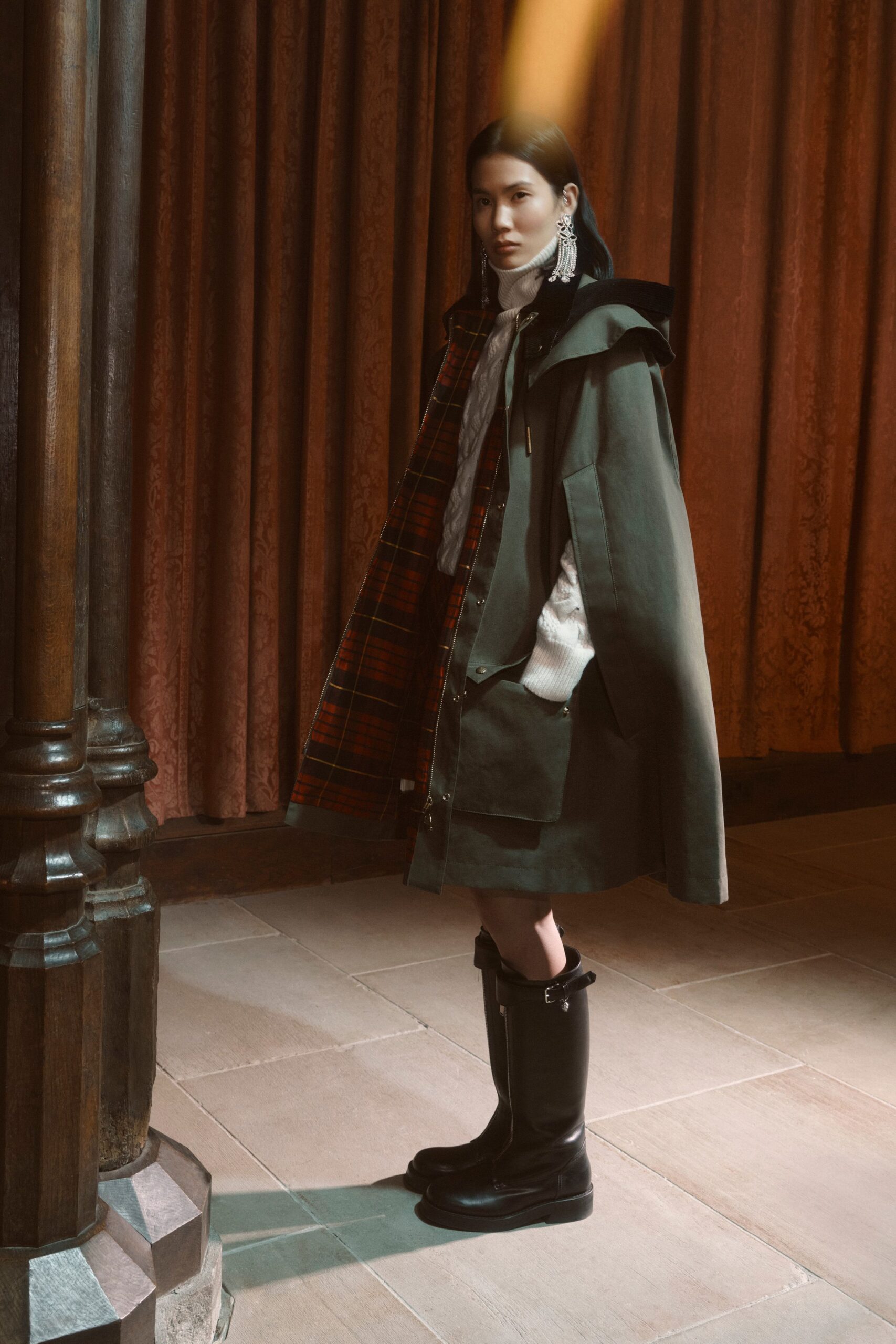 Dark olive cape coat, red tartan lining flashing inside the placket. White cable-knit turtleneck with a high ribbed neck. Matching olive mini with broad flap pockets. Crystal chandelier drops brushing the collar. Black riding boots with a strap crossing the ankle. Crossing a drafty manor corridor before dinner is called.