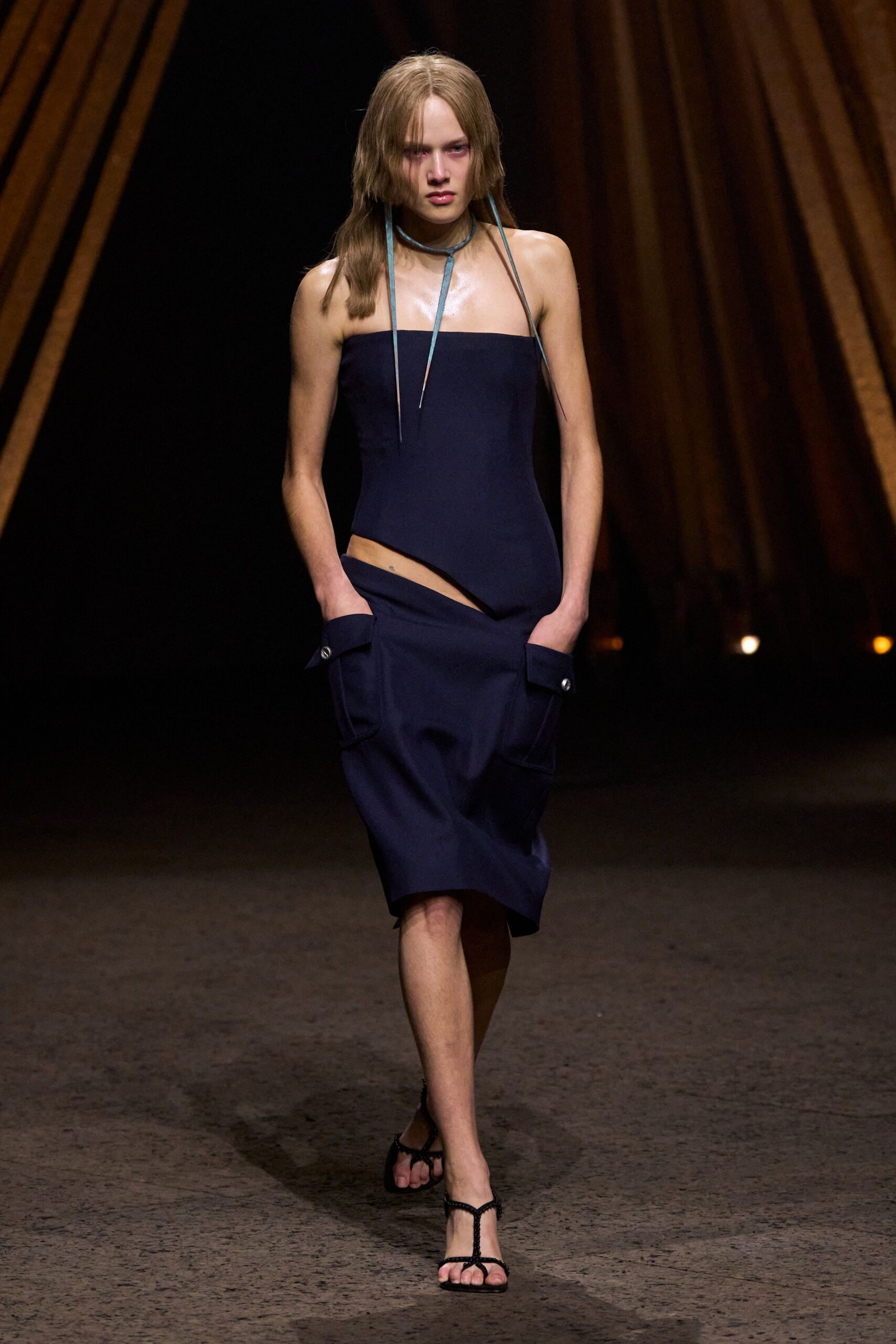 Powder-blue leather cords looped tight at the throat, metal tips hanging long. Ink column dress with a bandeau bodice and sharp diagonal waist cutout. Flap patch pockets jutting at the hips with a single silver snap. Black beaded T-strap sandals. Walking across a hotel courtyard after the pool has emptied out.