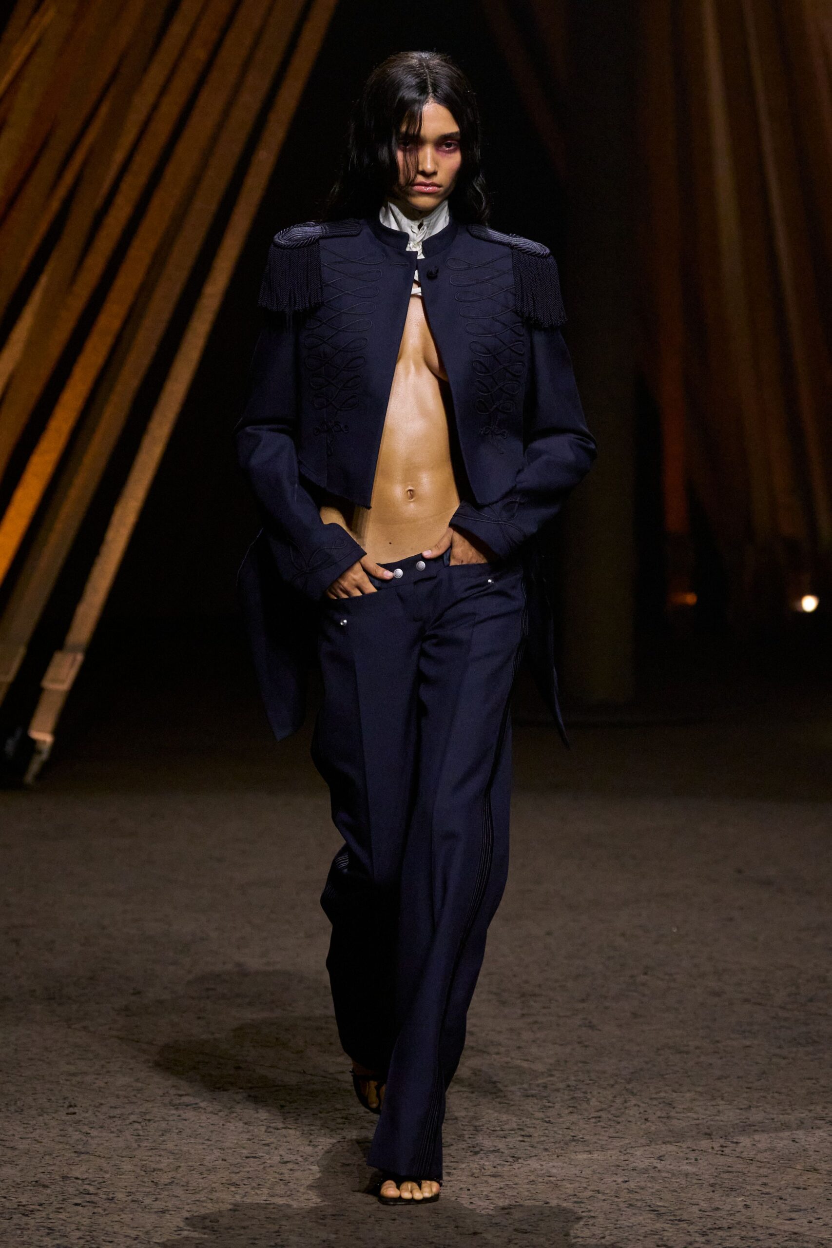 Ink-navy wool with braided frogging and heavy fringe epaulettes. White stand collar peeking through a tiny keyhole fastening. Low-slung navy trousers with pearl snaps glinting at the dropped waistband. Black barely-there sandals, toes fully exposed. Whatever you wear crossing a hotel lobby at dawn after losing the room key.