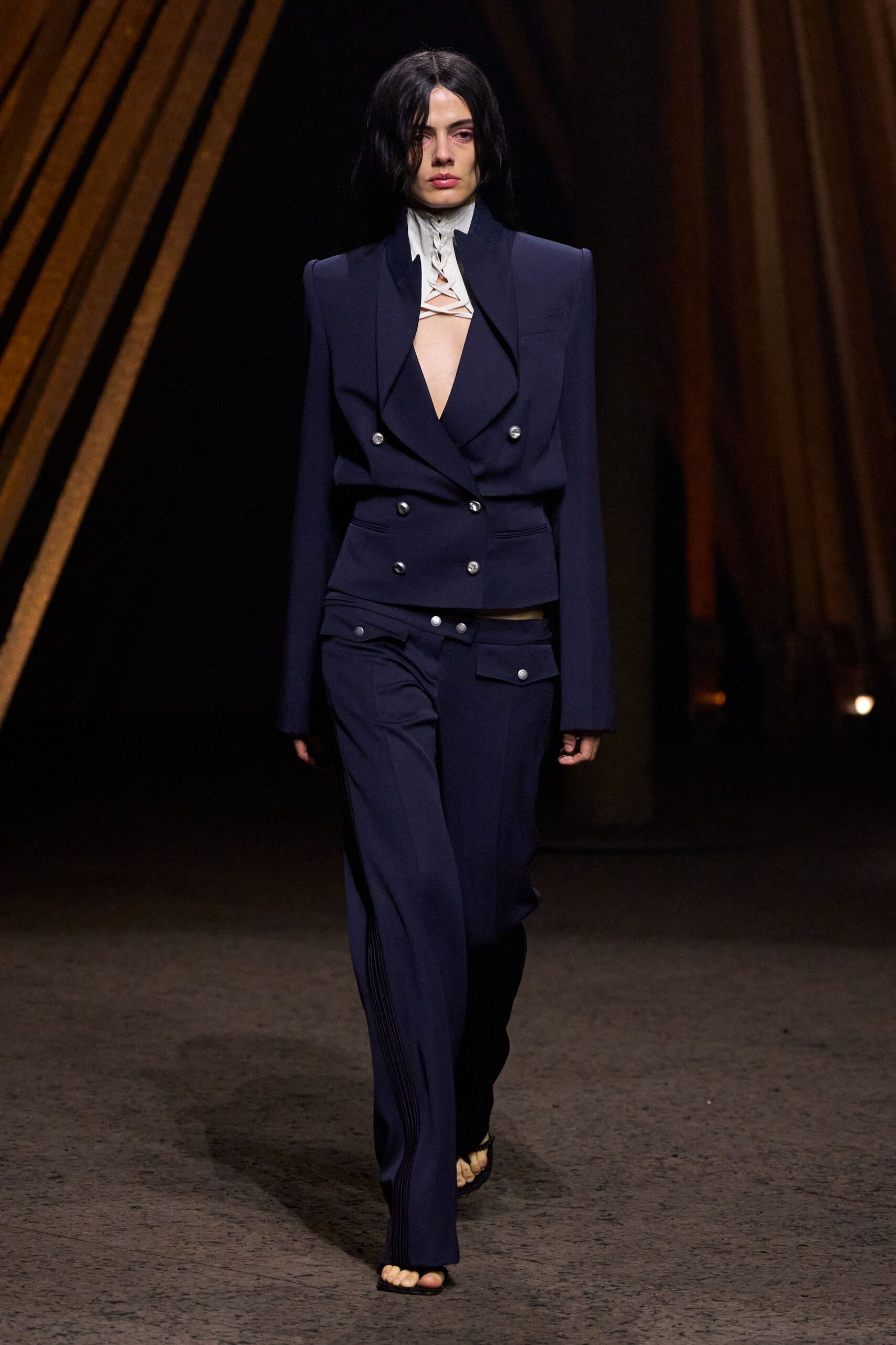 Ink navy blazer with sharp pagoda shoulders and silver crest buttons. Ivory funnel-neck underlayer laced open across the throat and chest. Low-slung navy trousers with flap pockets and pin-tucked side seams. Black thong sandals, toes pushed right to the edge. Walking out of a courthouse into wind that catches nothing.