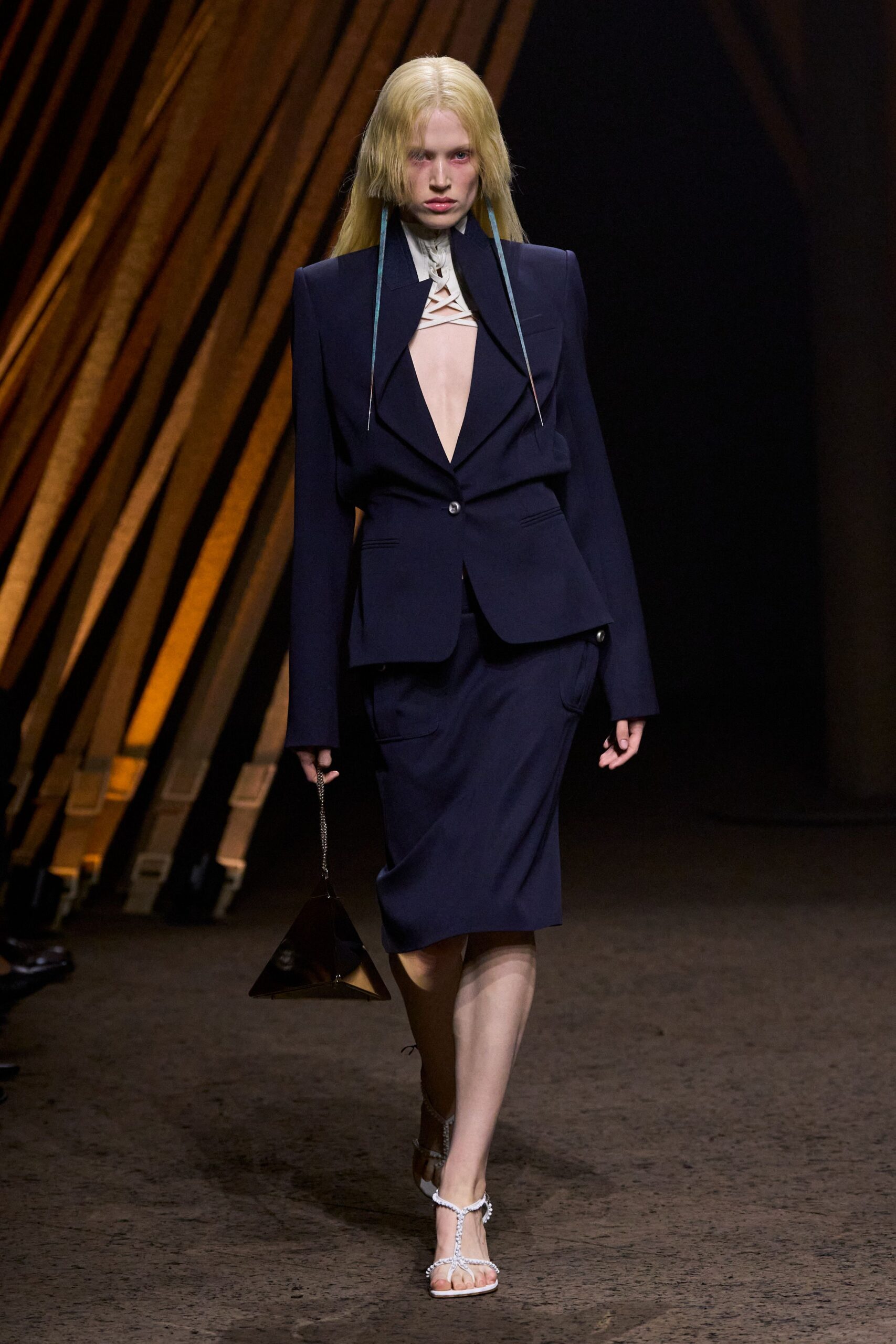 Inky tailored blazer cut into a plunging V, one low button. Ivory high throat with crisscross bands suspending the bare chest. Matching knee skirt with flap pockets slung low on the hips. Dark pyramid bag on a fine chain. White rope-strap sandals. After the lecture, still carrying the museum gift shop geometry.
