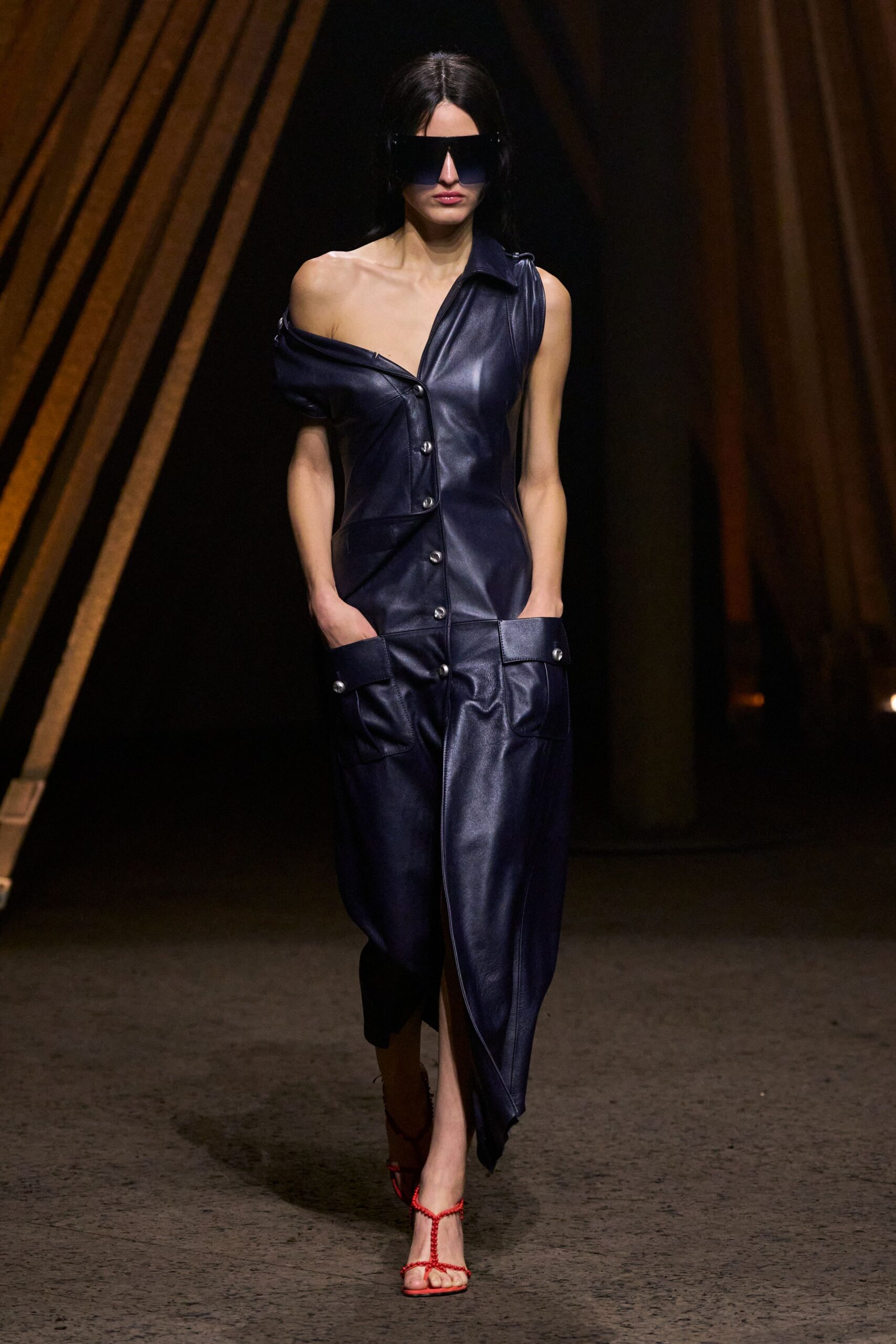 Oversized black shield visor with a hard straight brow. Ink-blue leather dress, one shoulder dropped, silver snaps marching off-center. Red cord T-strap sandals, thin ties biting across the toes. Crossing a hotel atrium after midnight with room keys and nowhere urgent.