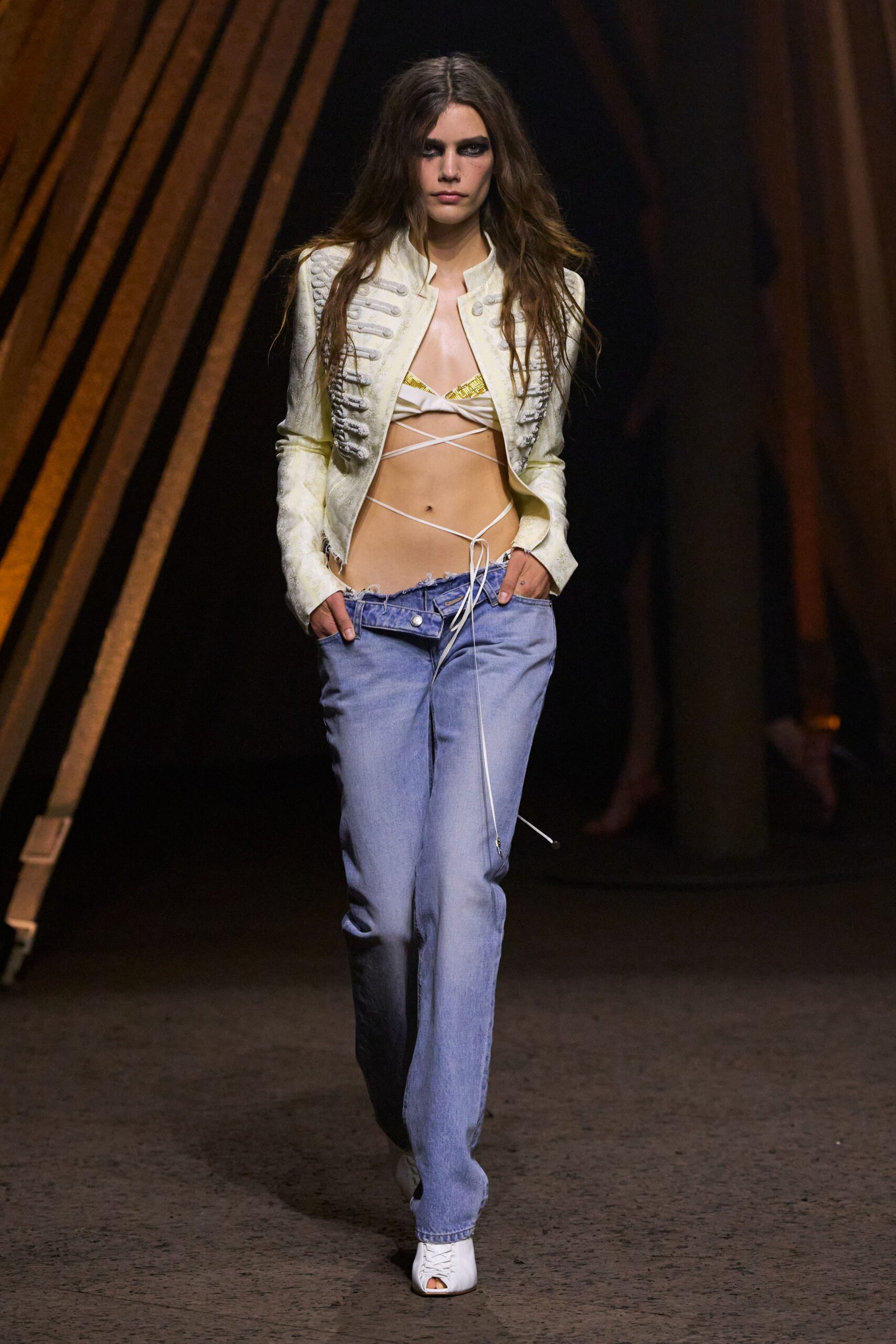 Ivory cropped jacket with loop-frog trim and frayed cuffs. Cream bra top with gold foil triangles and wraparound cords. Low-slung pale denim, waistband skewed open with ragged edge. White peep-toe mules. Whatever you wear back from the festival after sunrise.