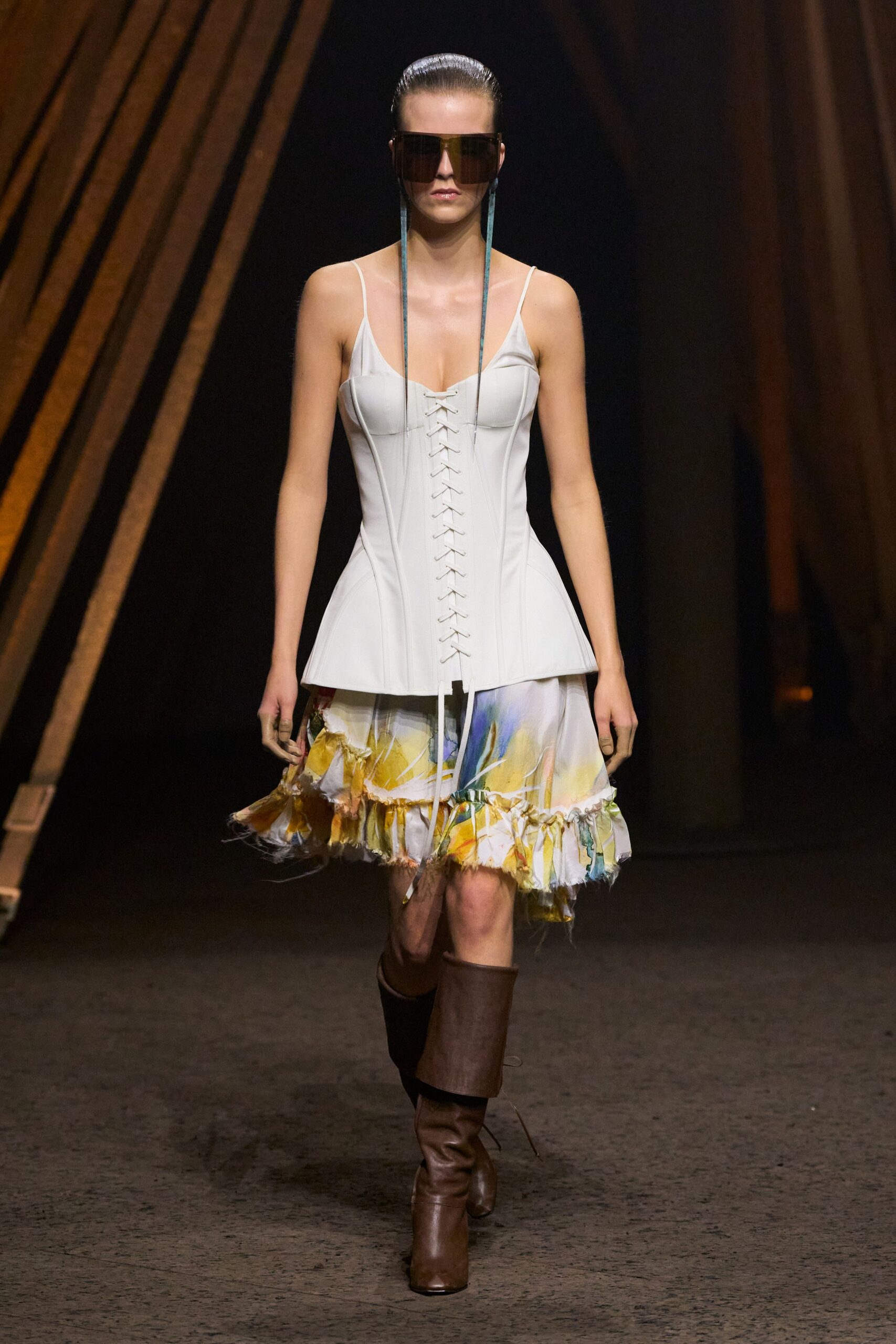 Oversized smoke shield lenses with long blue cords hanging past the bust. Pale corset bodice, center-laced, with thin straps and a peplum flare. Watercolor silk skirt, ruffled hem left fraying in loose threads. Brown leather boots with wide cuffed shafts. Walking out of a hotel atrium still carrying yesterday's bouquet.