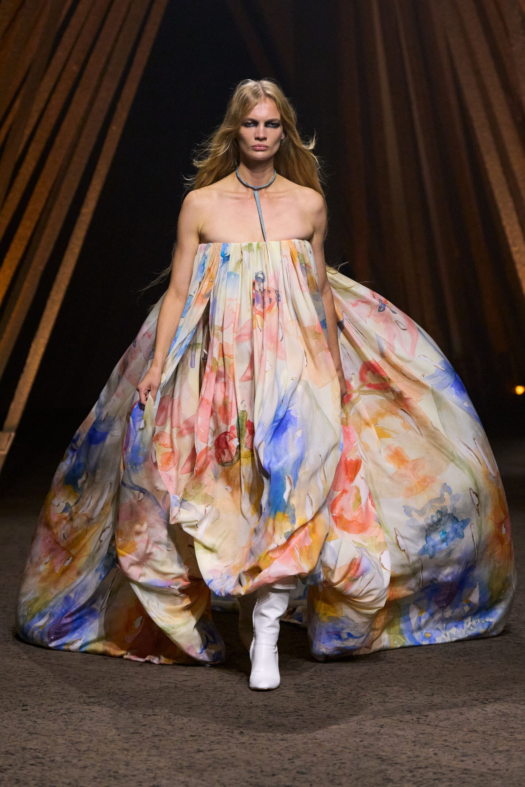 Powder-blue cord choker dropping into a single long front tie. Strapless balloon dress in watercolor floral silk, gathered hard across the bust. White knee boots peeking from the swollen hem. Crossing a museum atrium with an armful of fresh posters.