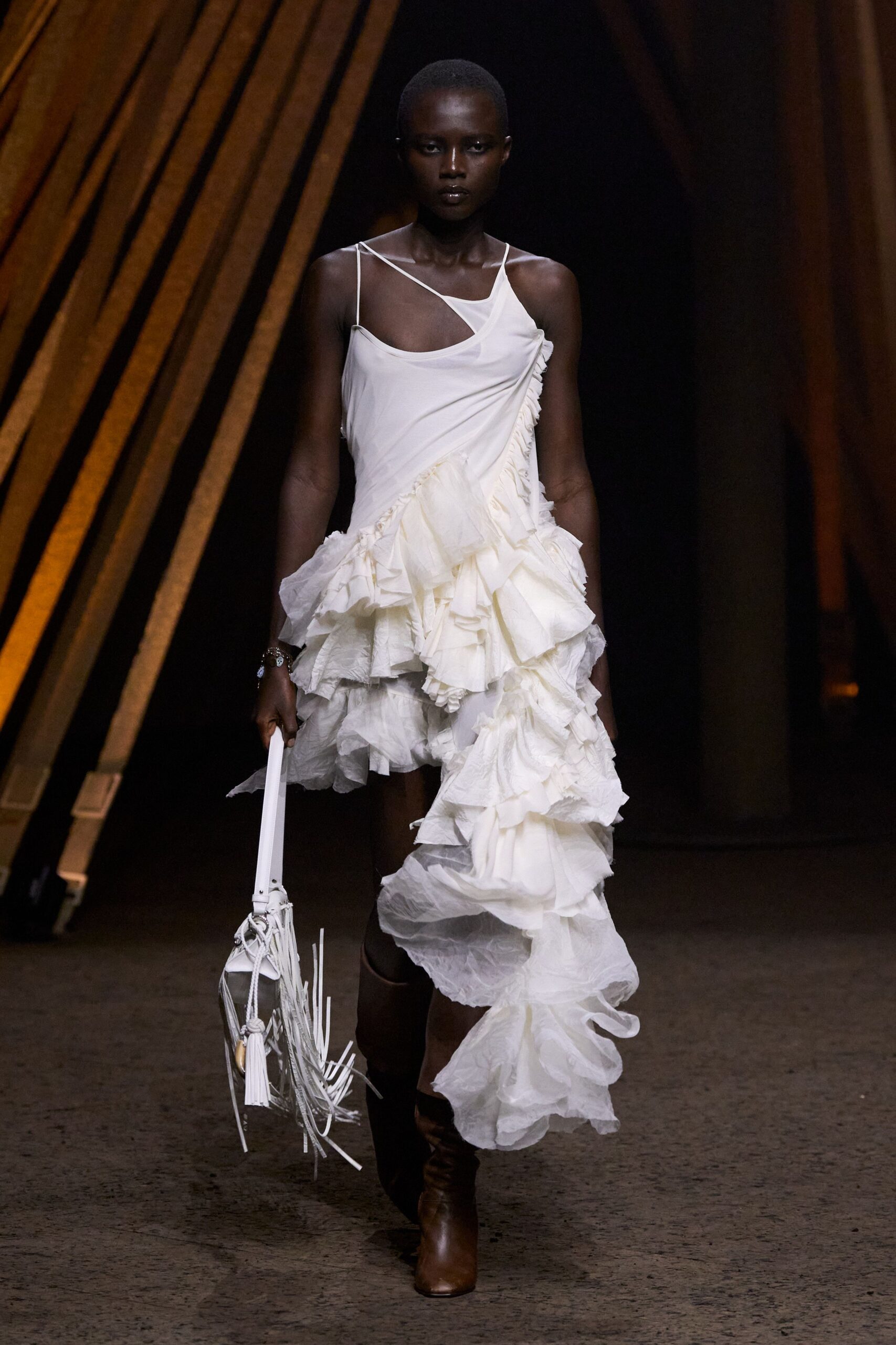 Ivory one-shoulder dress in airy jersey and paper-light ruffles, one strap crossing the bare chest. White bucket bag with long fringe and a tassel swinging past the handle. Chestnut knee boots in smooth leather. Walking across a hotel courtyard after midnight with flowers already collapsing.
