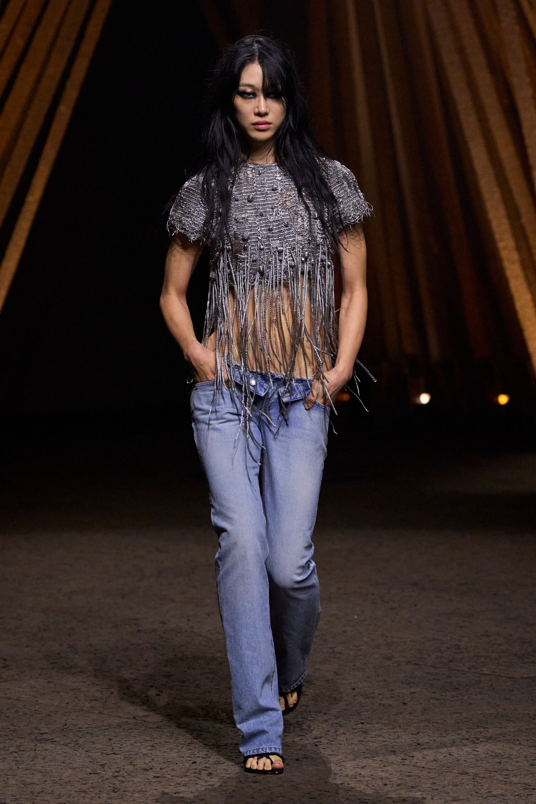 Silver boucle crop top with bobbles and long dangling fringe. Pale-wash jeans slung low, waistband twisted under the navel. Black thong sandals. Walking out of a warehouse show into damp midnight heat.