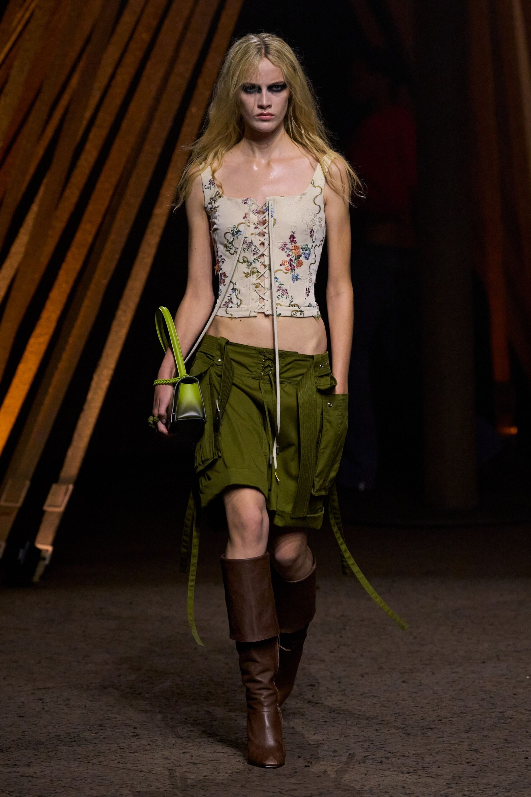 Cream floral corset top, front lacing left hanging past the hem. Olive cargo mini with low lace-up waist and long webbing straps. Acid-green shoulder bag with a braided handle edge. Brown leather knee boots, softly slouched at the ankle. Walking back from the garden center with dirt under one thumbnail.