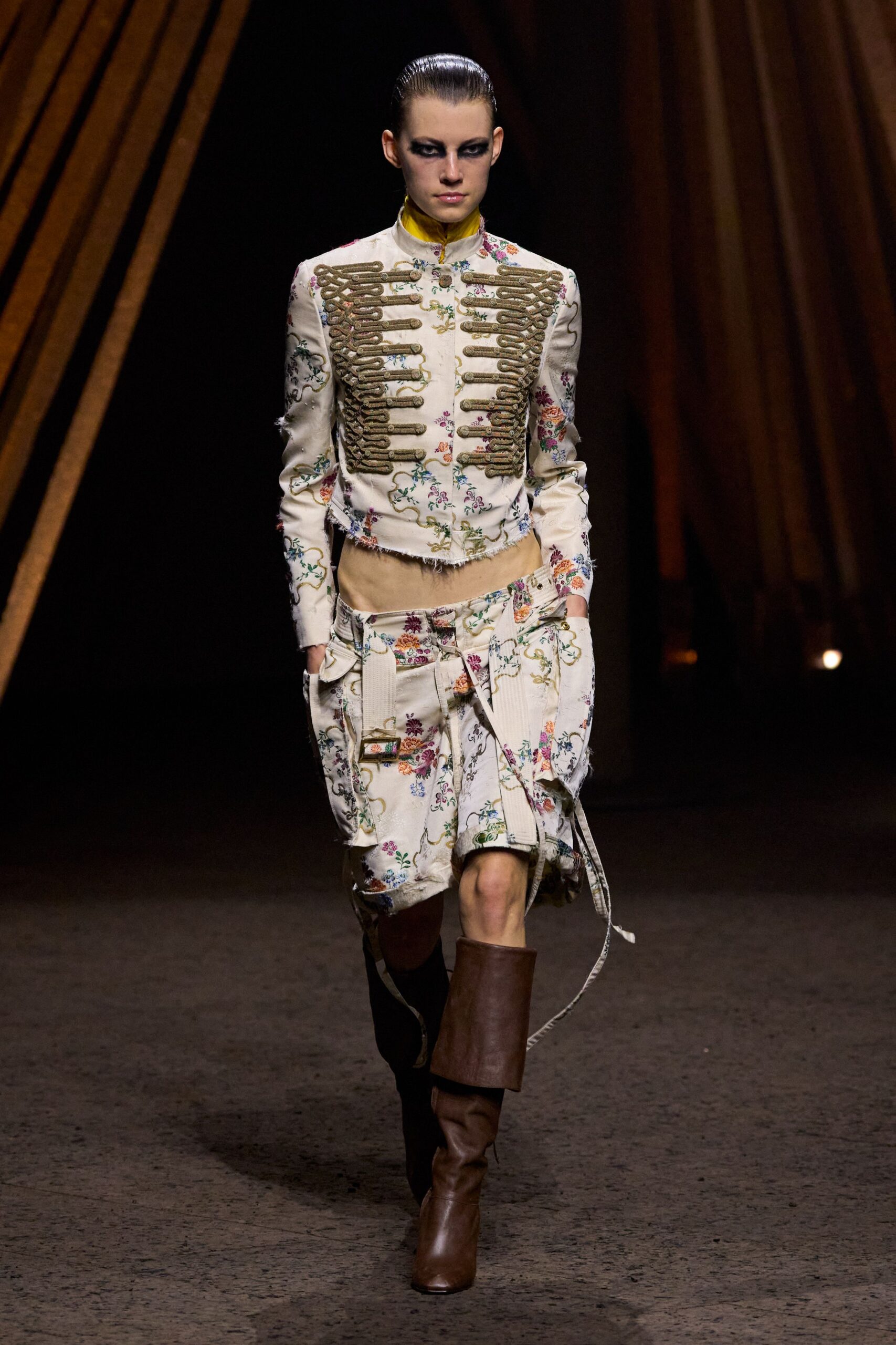 Cream floral jacket with bullion frogging, frayed hem and cuffs. High saffron stand collar flashing above the neckline. Matching floral cargo shorts slung low, with hanging webbing straps. Chestnut leather boots, one shaft folded thick at mid-calf. For crossing the fairground after rain, pockets full and nowhere urgent.