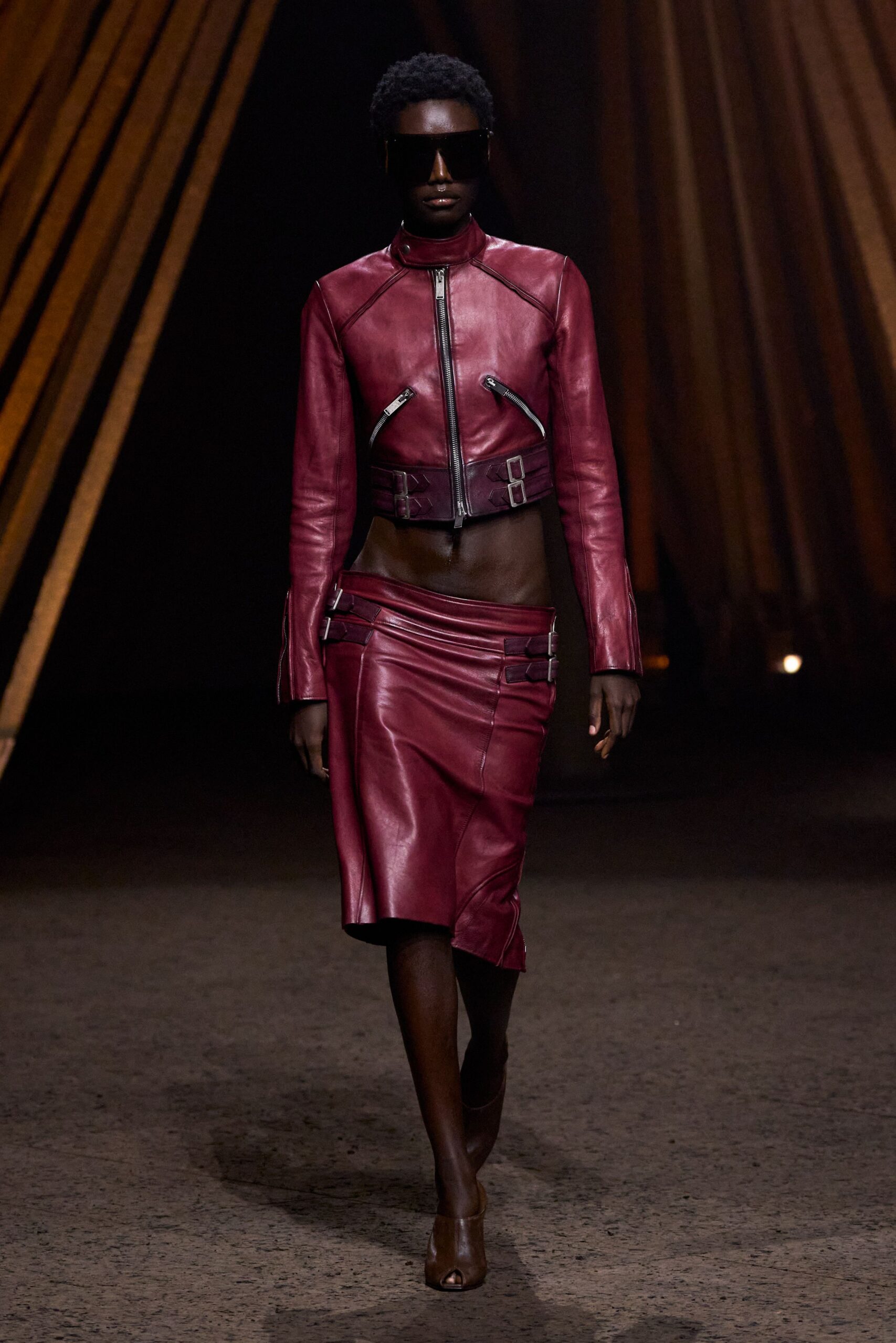 Oversized black shield visor. Bordeaux leather biker jacket, cropped above the navel with buckled waist tabs. Matching low-slung leather pencil skirt, wrapped seams and side buckle straps. Brown peep-toe ankle boots. Whatever you wear to cross a hotel lobby at sunrise unnoticed.