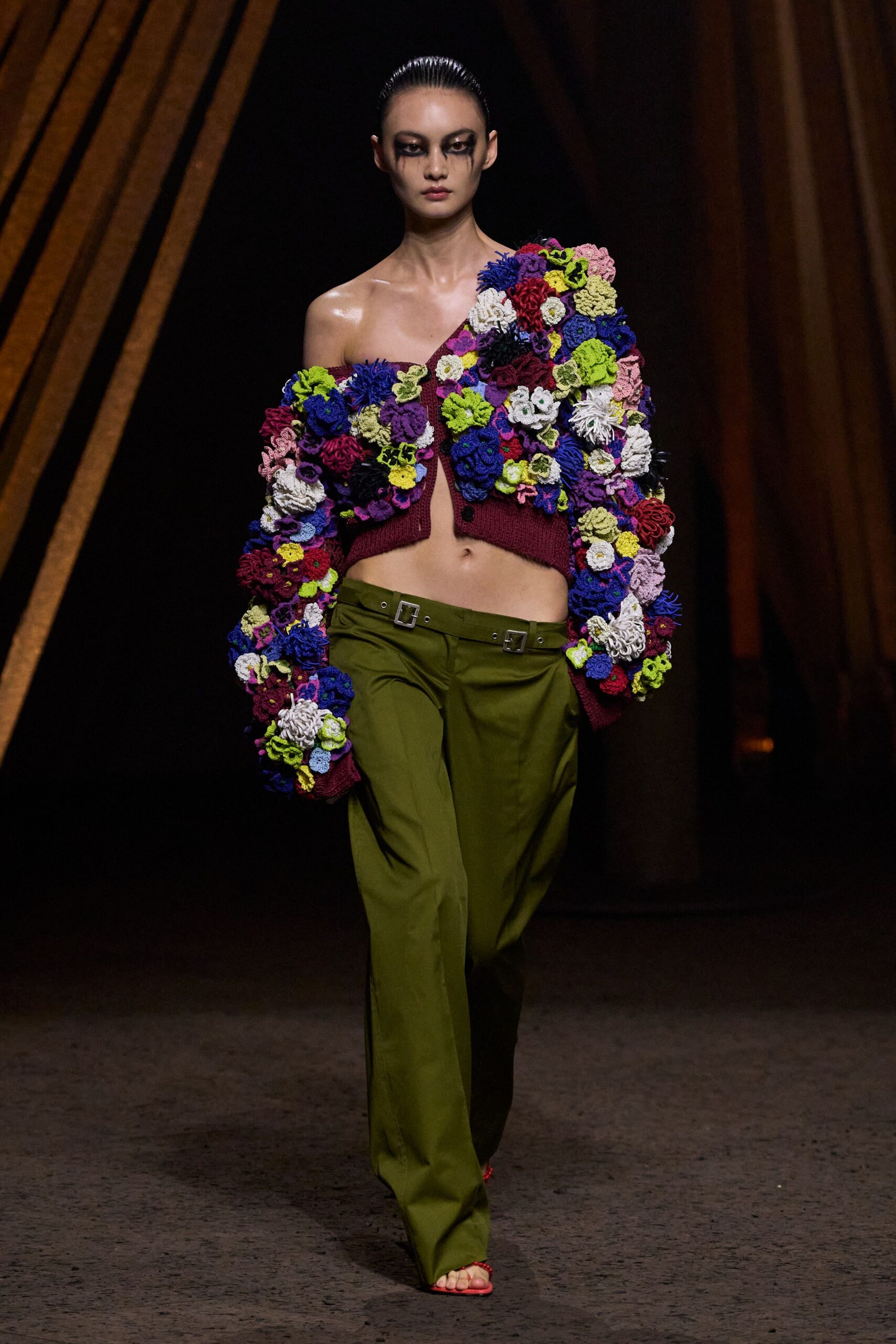Burgundy knit cardigan crusted with dense crocheted flowers, one shoulder dropped bare. Olive tailored trousers slung low with twin square-buckle waistband straps. Coral thong sandals, barely peeking from the pooling hem. Walking through a botanic conservatory after dark, still smelling faintly of wet soil.