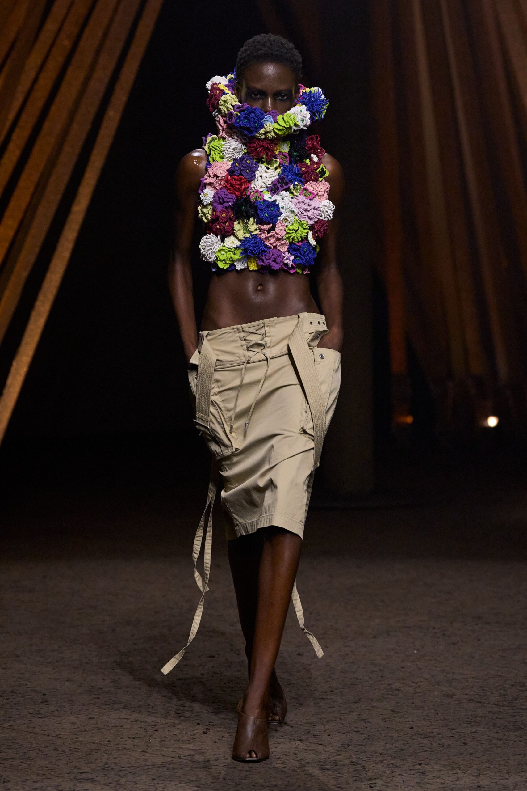 Sleeveless cropped top built from dense multicolour crochet flowers, rising into a face-framing ruff. Sand utility wrap skirt in crisp twill, laced low with long belt ends swinging. Brown leather peep-toe mules. Walking under timber rafters with sawdust still in the air.