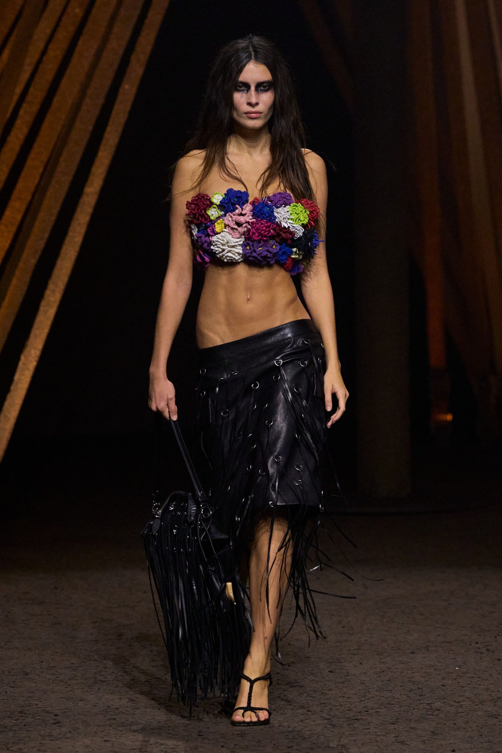 Strapless bustier built from dense crocheted rosettes in acid-bright colours. Black leather wrap skirt slung low, ring-laced strips whipping past the knee. Long black fringe bag dragging almost to the floor. Barely-there black T-strap sandals. Crossing a fairground after rain, prizes and power cables underfoot.