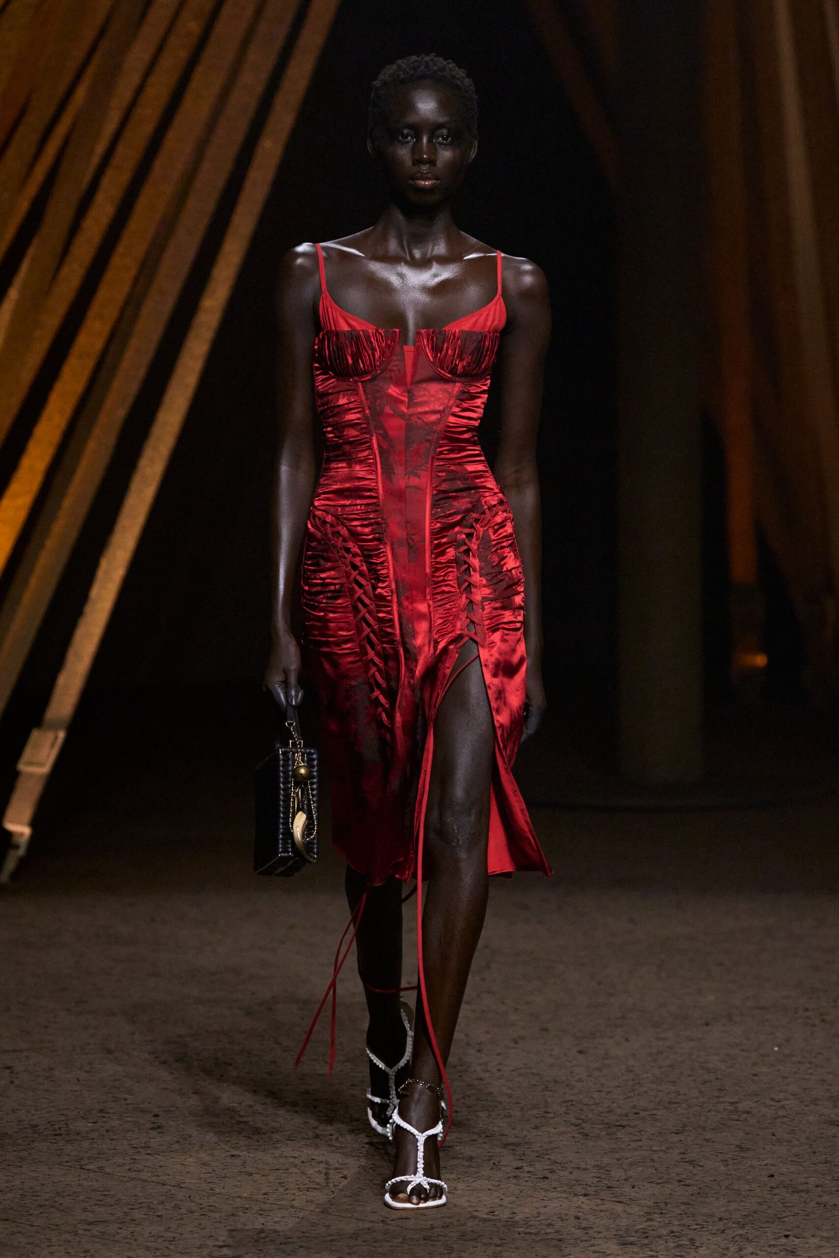 Crimson satin slip dress, ruched underwired cups and corset-laced hips. Black box bag with a tusk charm swinging from the clasp. White beaded thong sandals, ankle chain catching above the instep. For arriving at a terrace wedding just after the storm broke.