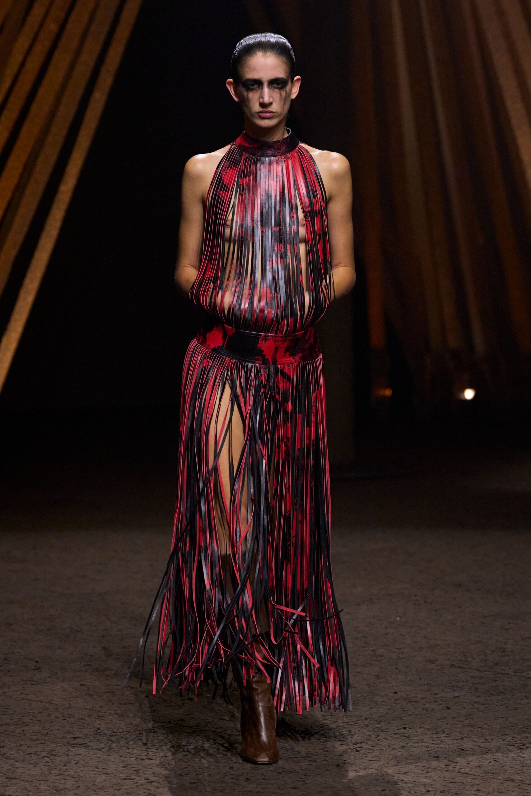 High-neck red-and-black print dress sliced into long leatherlike strips over the torso. Brown ankle boots with a blunt square toe. Crossing a fairground after rain, with ash smudged under both eyes.