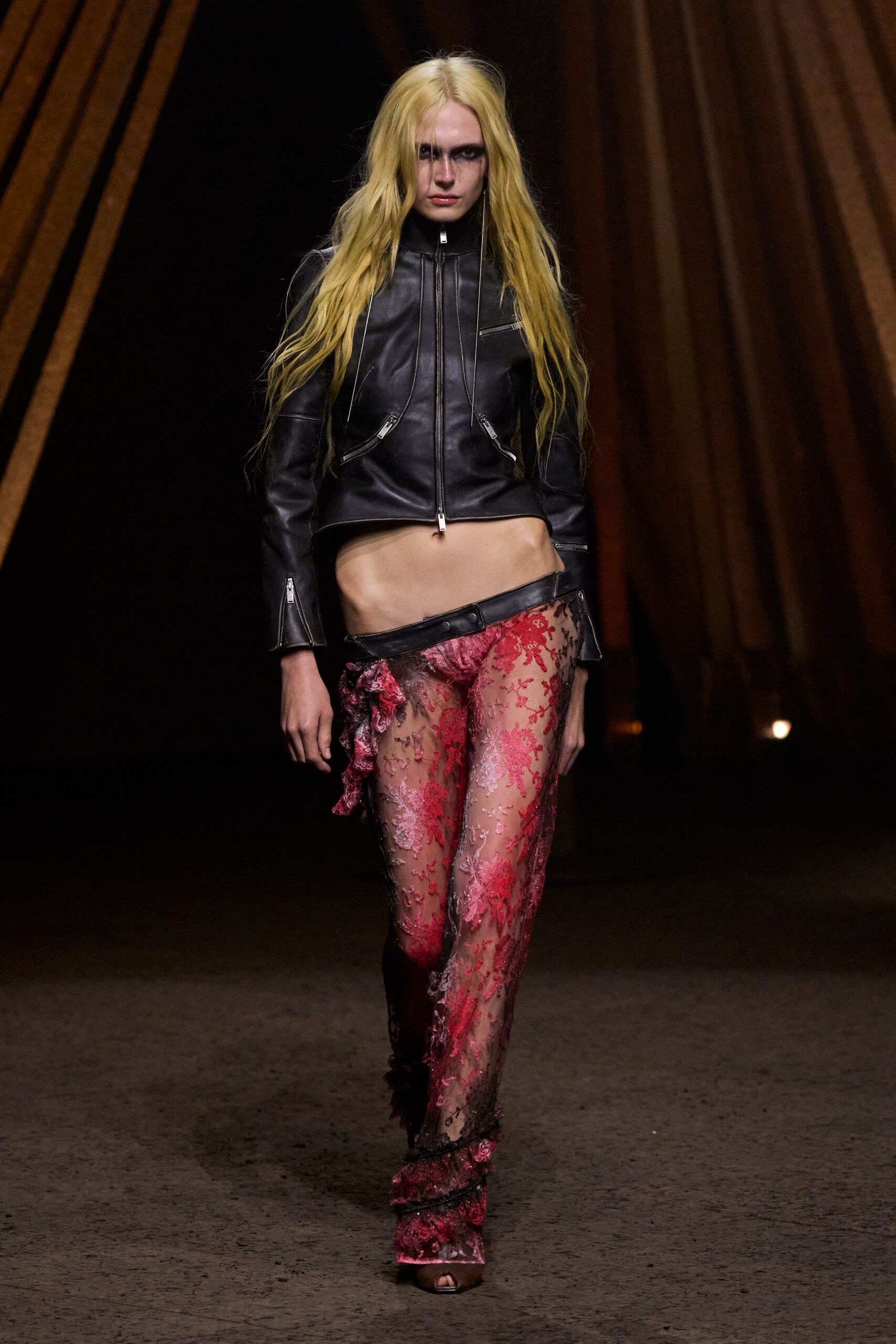 Black leather moto jacket, cropped hard with silver zips at chest and cuffs. Sheer mesh trousers crusted with pink corded lace, waistband dropped off one hip. Peep-toe platform heels veiled by the trailing lace hem. Whatever you wear home after dawn when the club carpet is still damp.