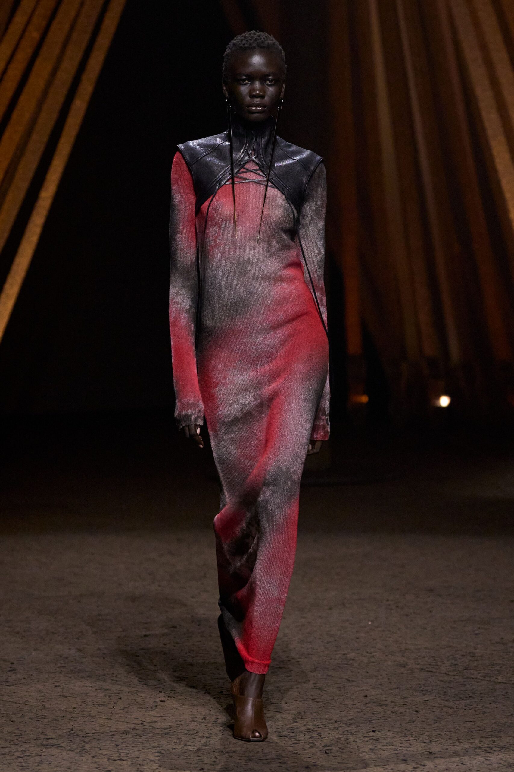 Black leather shoulder cape, cut high with laced cords crossing the chest. Fine ribbed column dress airbrushed from soot grey into bruised crimson. Brown leather ankle boots with a peep toe slit. Crossing a fairground after rain, generators still humming in the dark.