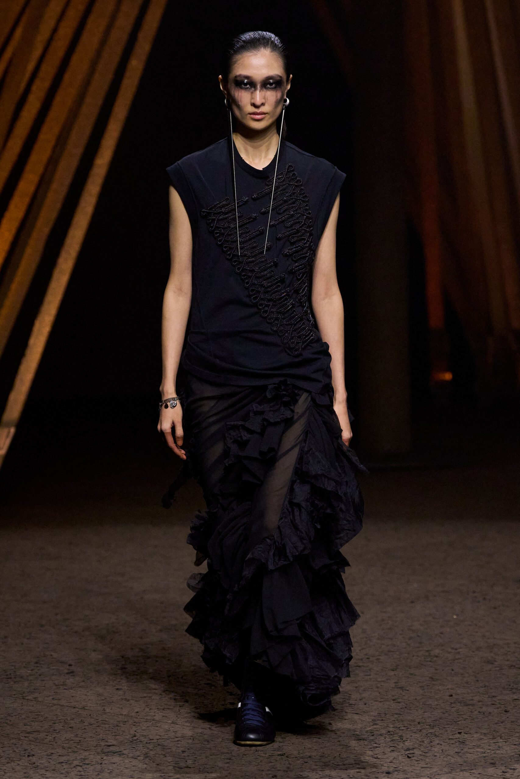 Black sleeveless jersey top with dense soutache looping diagonally across the chest. Sheer black skirt wrapped over the hips, exploding into crinkled ruffles. Long silver drop earrings. Charm bracelet at one wrist. Black trainers with a white toe edge. Walking through an empty opera house after the cleaning crew left.