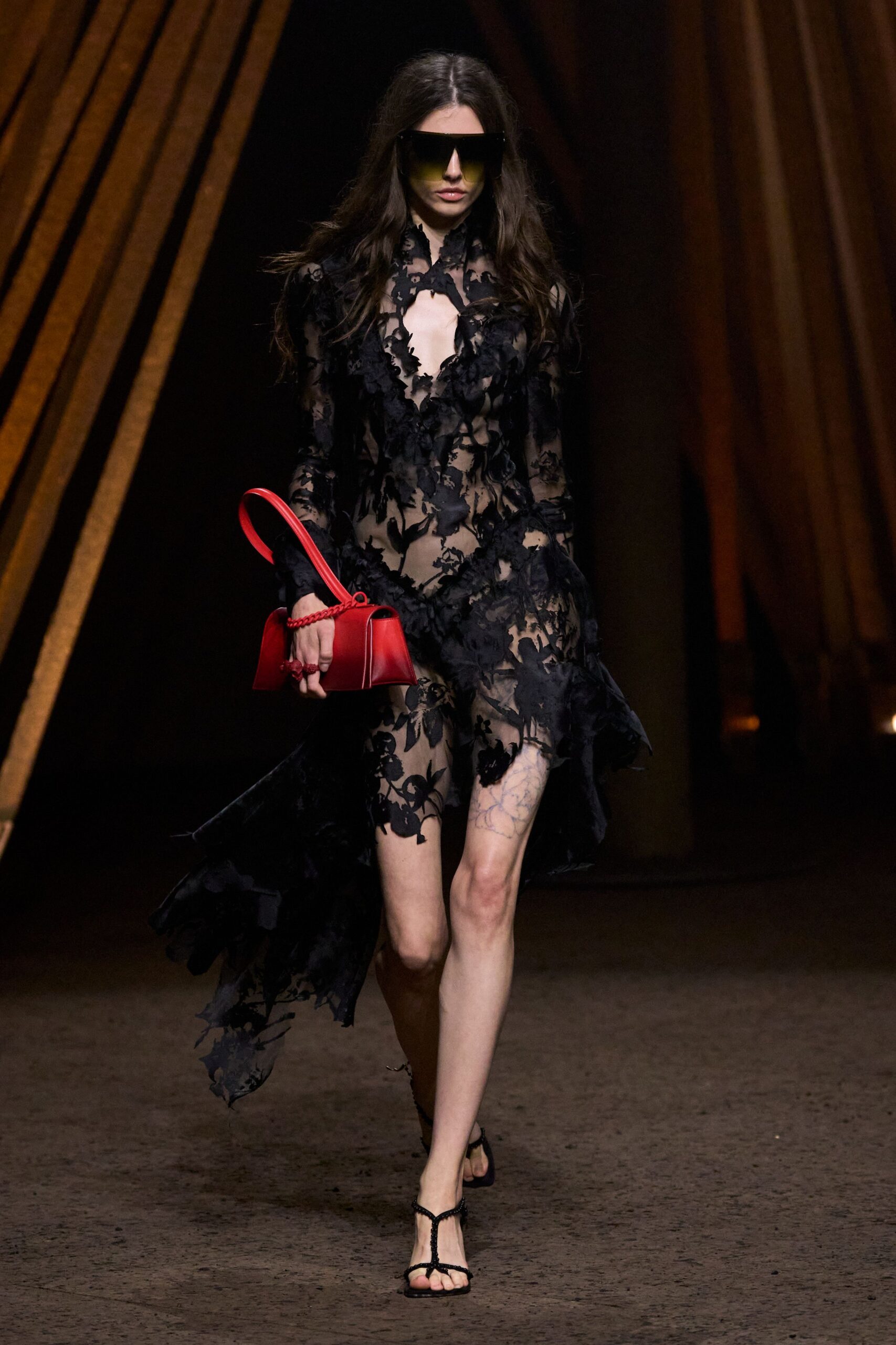 Oversized shield sunglasses fading from smoke to acid olive. Sheer black lace dress with appliqued petals and a trailing asymmetric hem. Red top-handle bag with a braided strap looped over the flap. Black thong sandals with slim crystal-studded T-straps. Crossing a hotel atrium after midnight with the flowers already crushed.