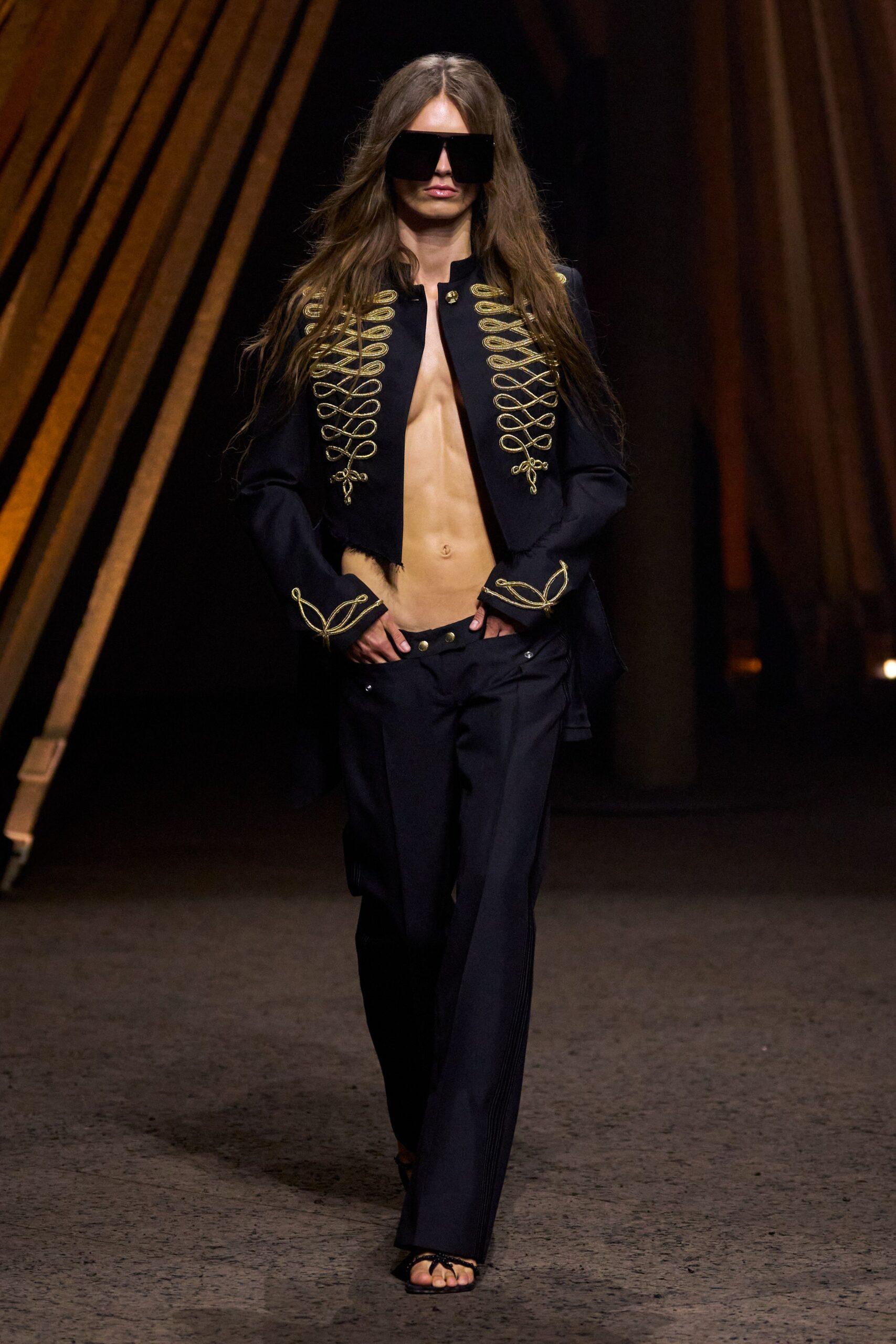 Oversized black visor lenses hiding half the face. Black military jacket traced with gold cord frogs, hem left raw. Low-slung black trousers with brass-tab waistband hanging open. Black thong sandals with a knotted tie across the toes. For ducking out the stage door while the brass section is still packing up.